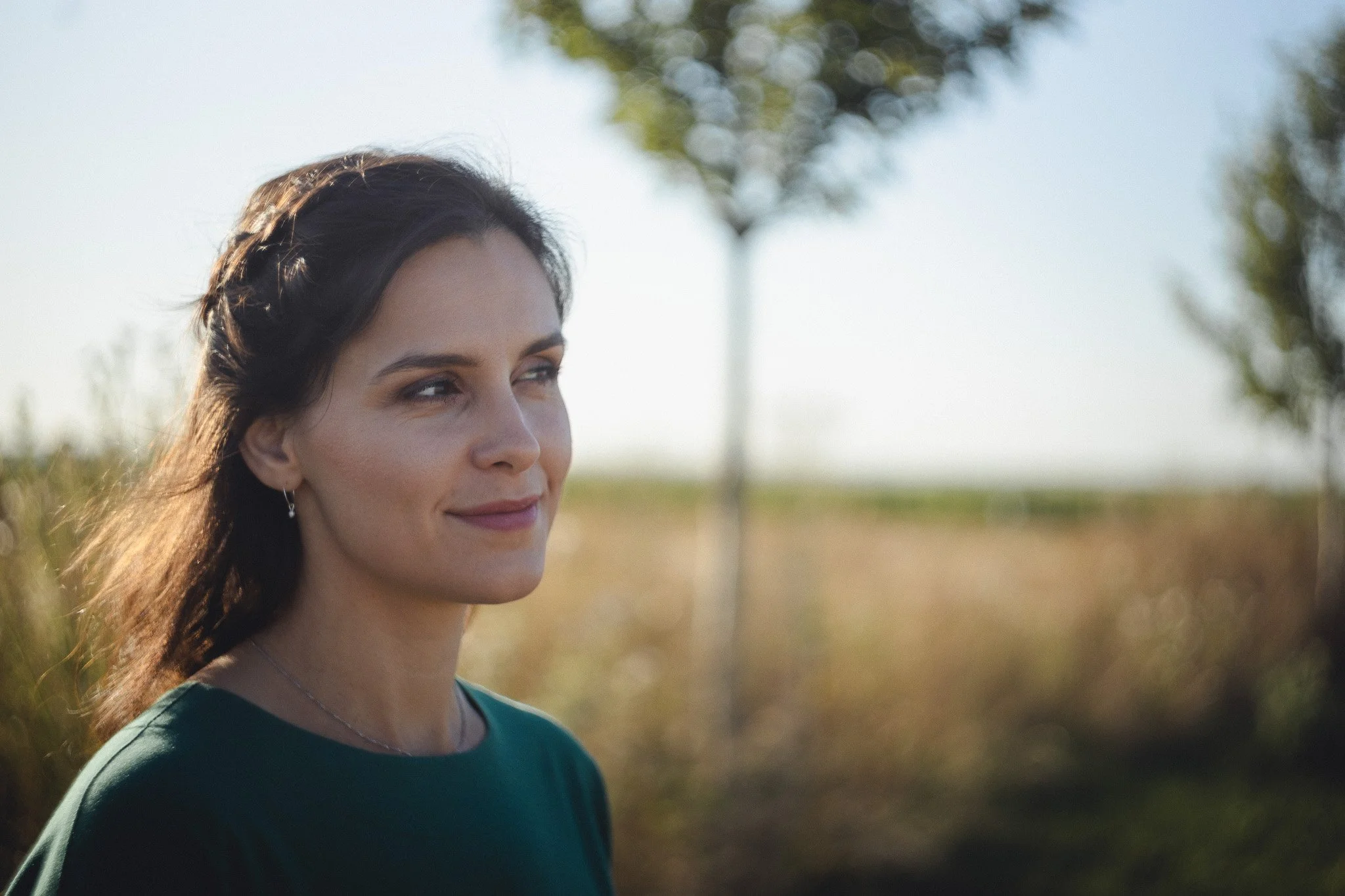 A woman with dark hair and earrings outdoors on a sunny day with trees and a field in the background