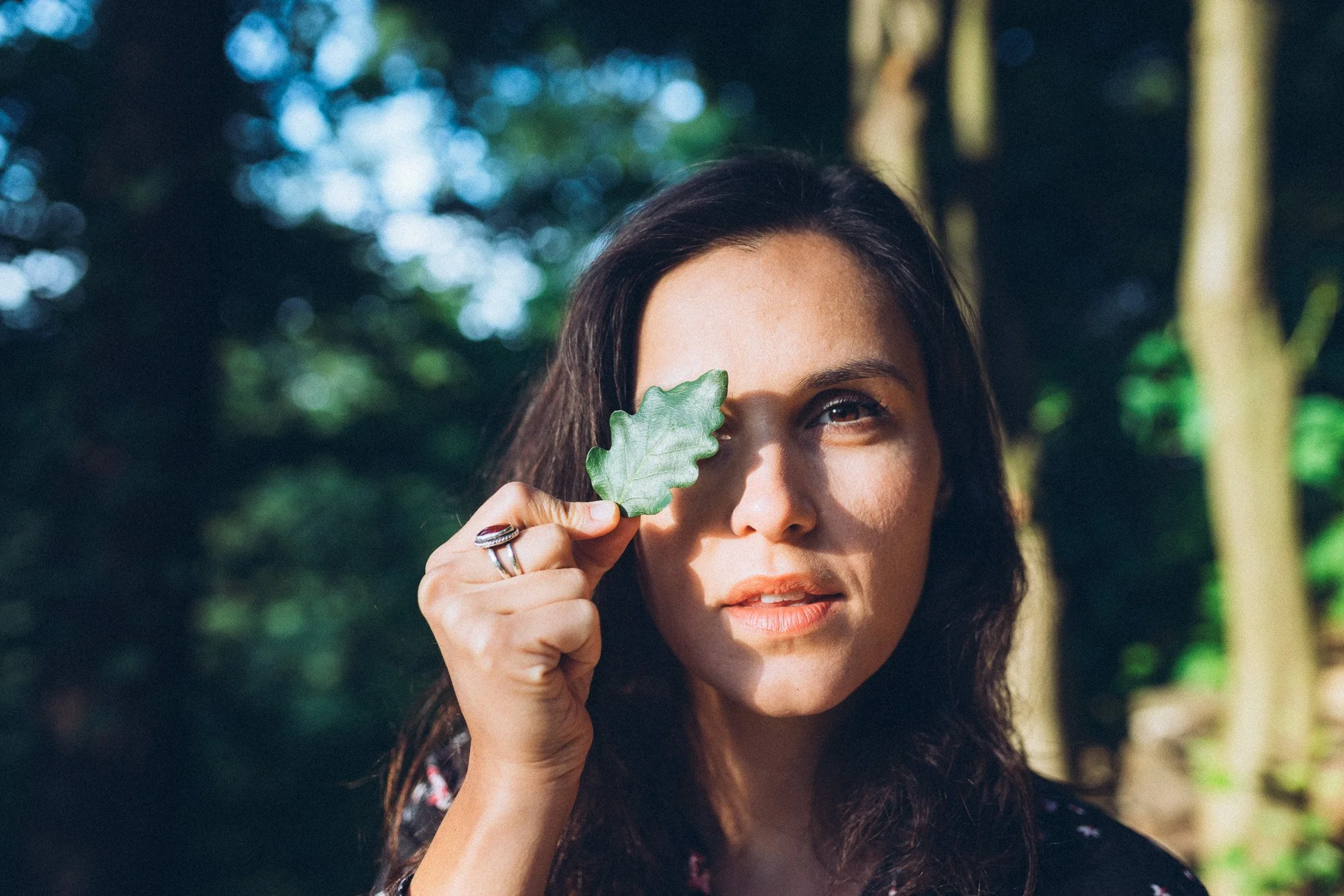 A woman with wavy dark hair holds a green leaf up to her eye while standing outdoors surrounded by trees and sunlight.