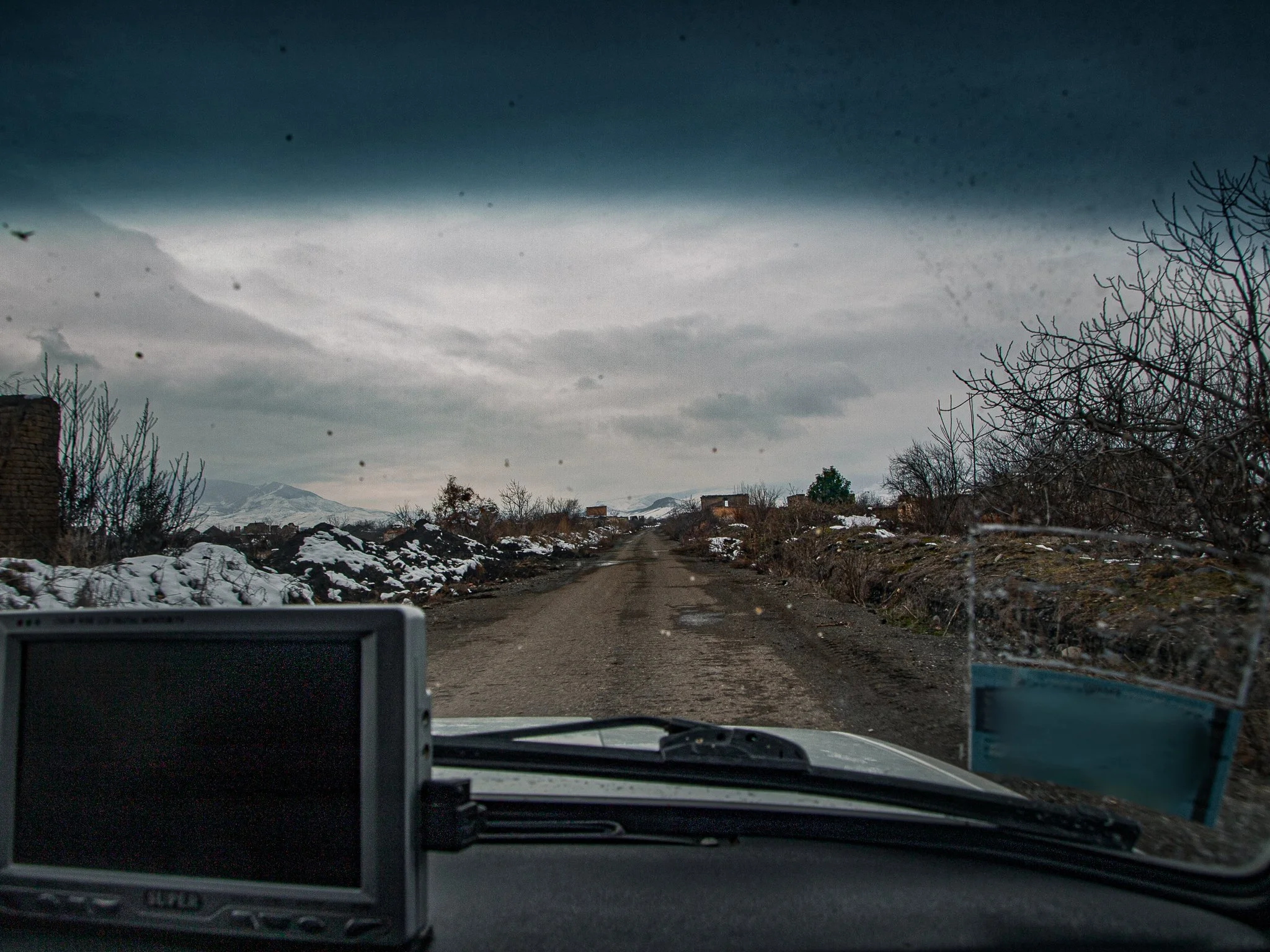 Unpaved dirt road in a rural area with snow on the ground and leafless bushes on either side, under a cloudy sky.