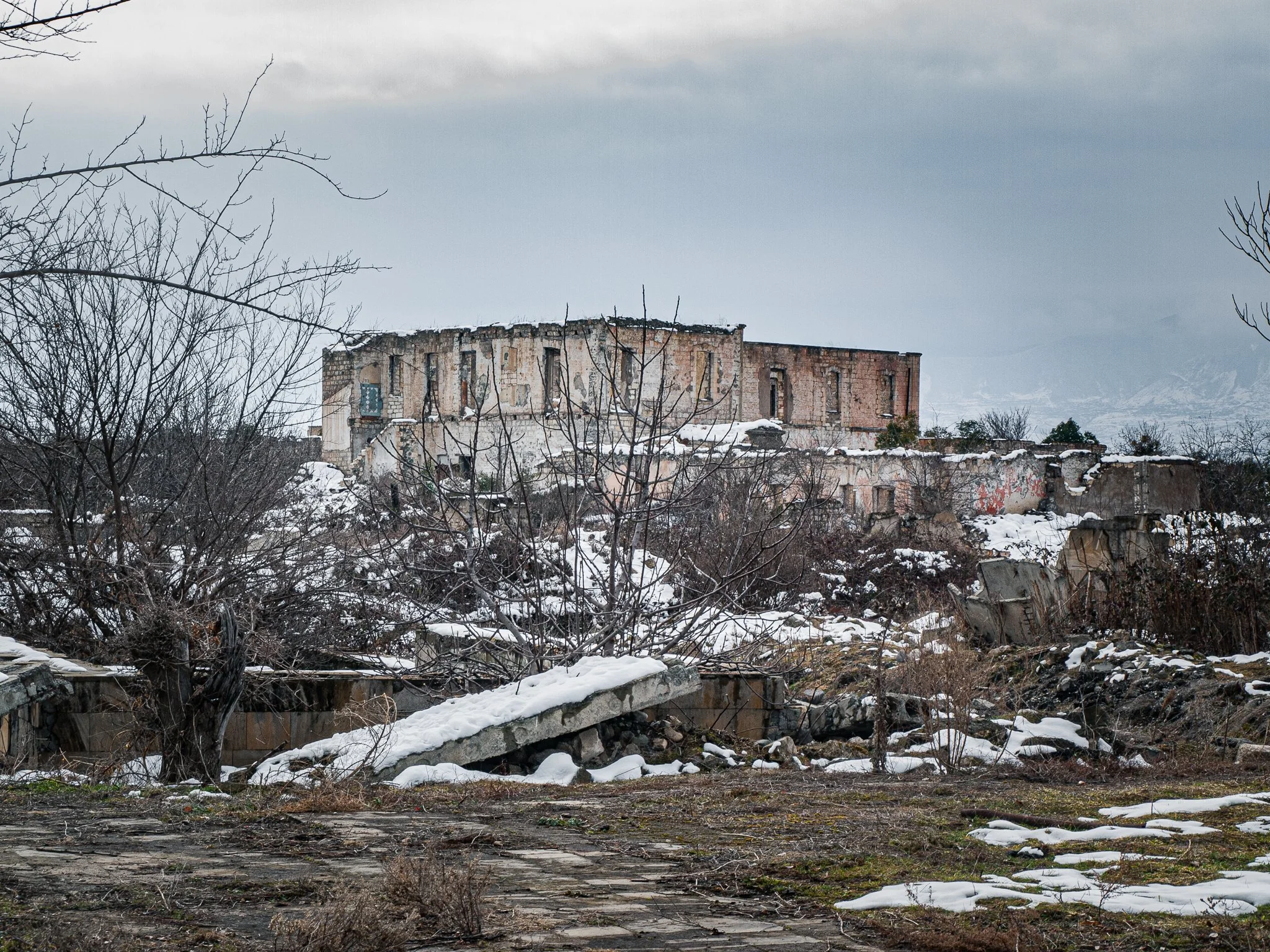 Abandoned, dilapidated building on a snowy hillside, with leafless trees and gray sky.