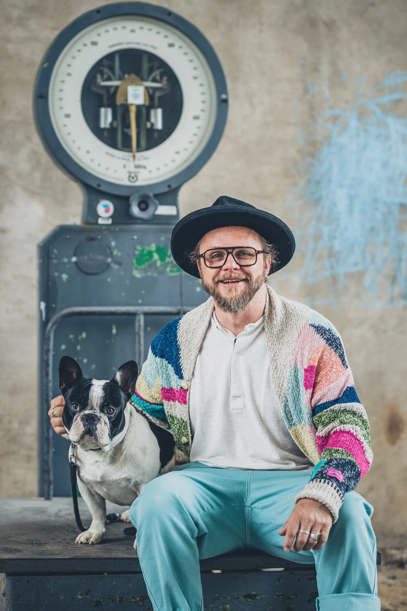 A man with glasses, a beard, and a black hat sitting on a bench with a French bulldog, wearing a colorful knit cardigan and light blue pants, in front of a vintage clock on a concrete wall.