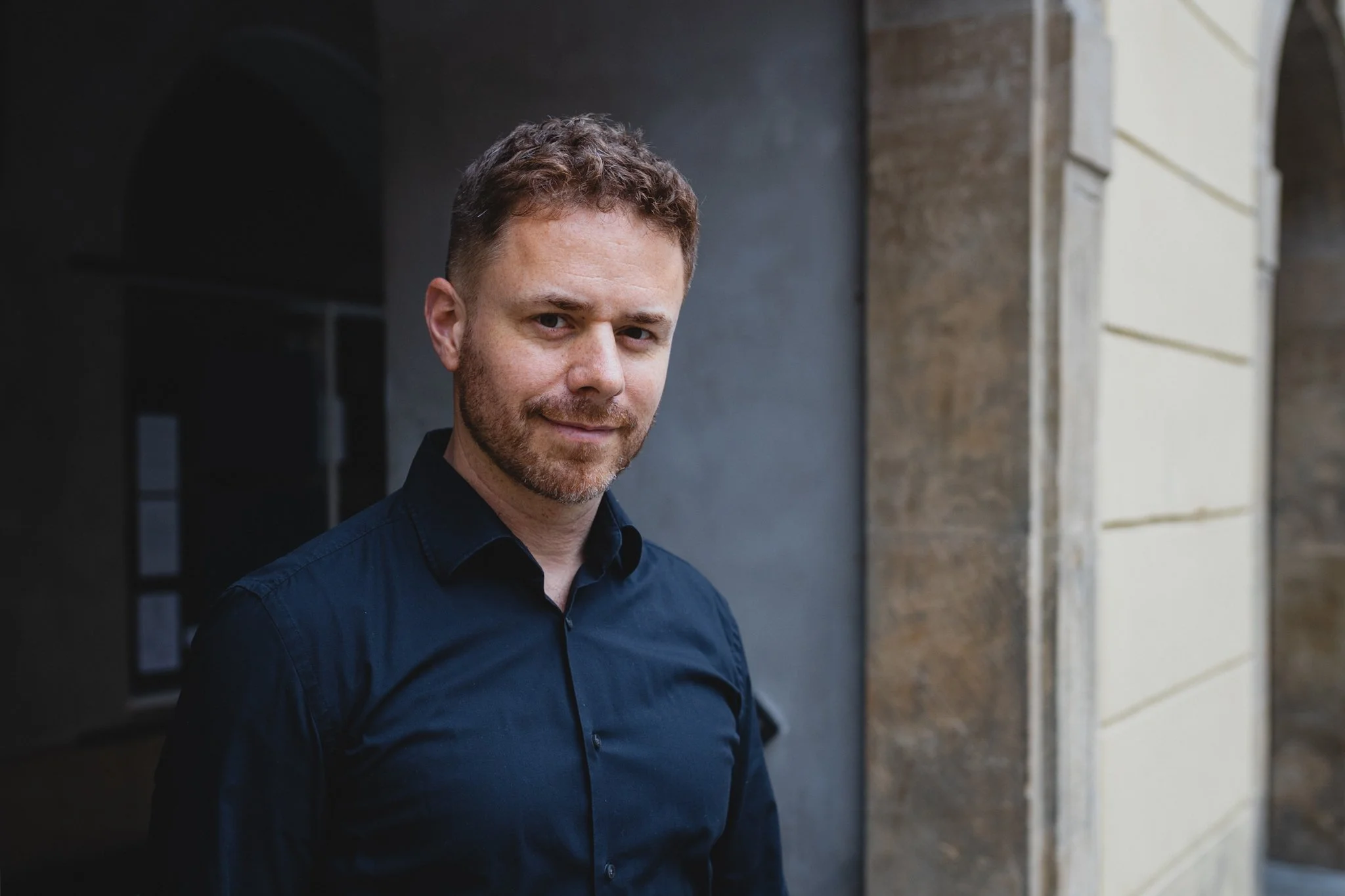 A man with light brown hair and a beard wearing a dark blue shirt standing outdoors near a stone building.