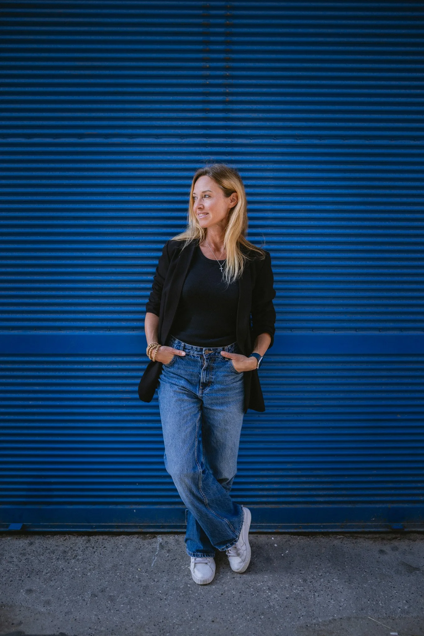Woman with blonde hair wearing a black jacket, black shirt, blue jeans, and white sneakers standing against a blue metal wall.