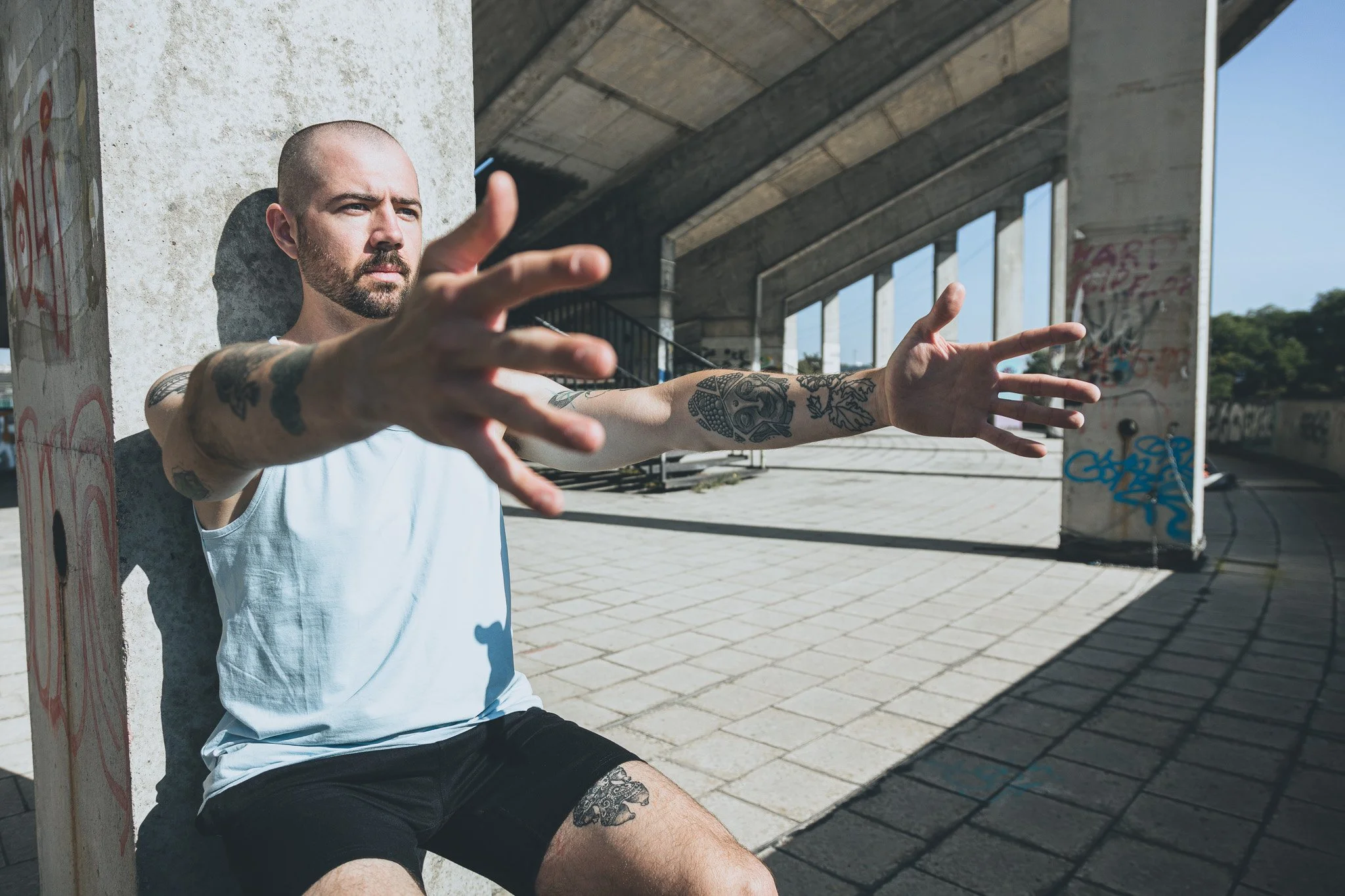 A man with tattoos on his arms and legs, wearing a white tank top and black shorts, sits under a concrete bridge taking a selfie with his arms extended forward.