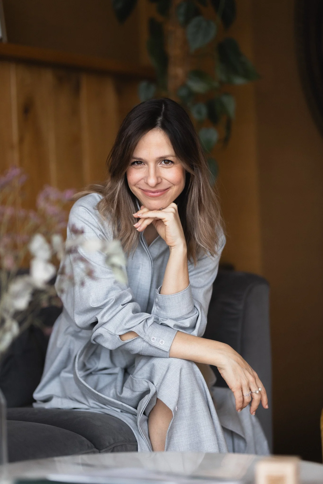 A woman with shoulder-length brown hair and light makeup smiling, sitting on a dark gray couch in a cozy room with wooden walls and green plants, dressed in a light gray casual top.