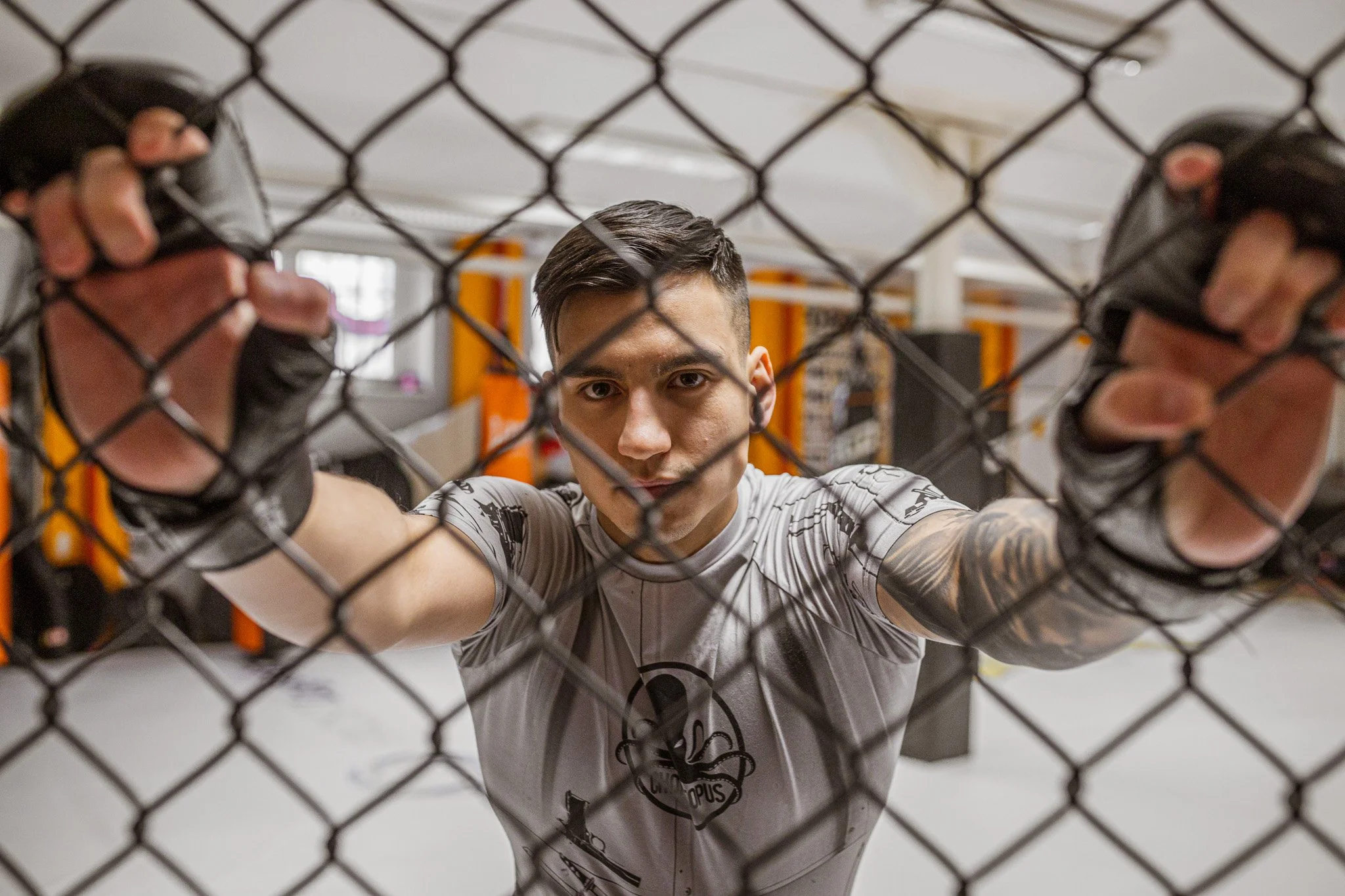 A young male mixed martial arts fighter looking through a chain-link fence, wearing a gray rash guard with a logo, inside a gym with orange equipment in the background.
