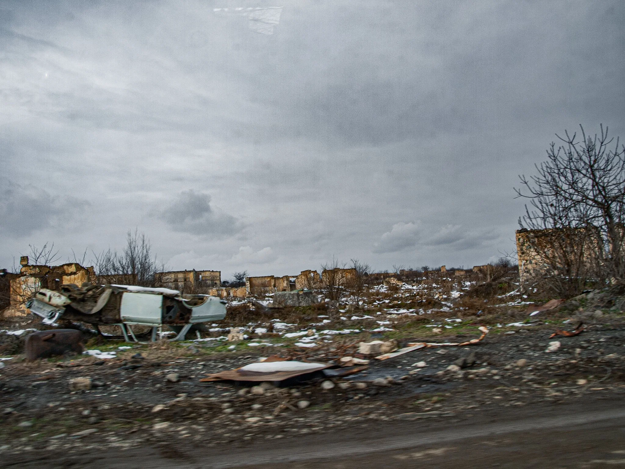 A scene of destruction and debris with abandoned, damaged buildings and a burned-out car in a barren, snowy landscape under a cloudy gray sky.