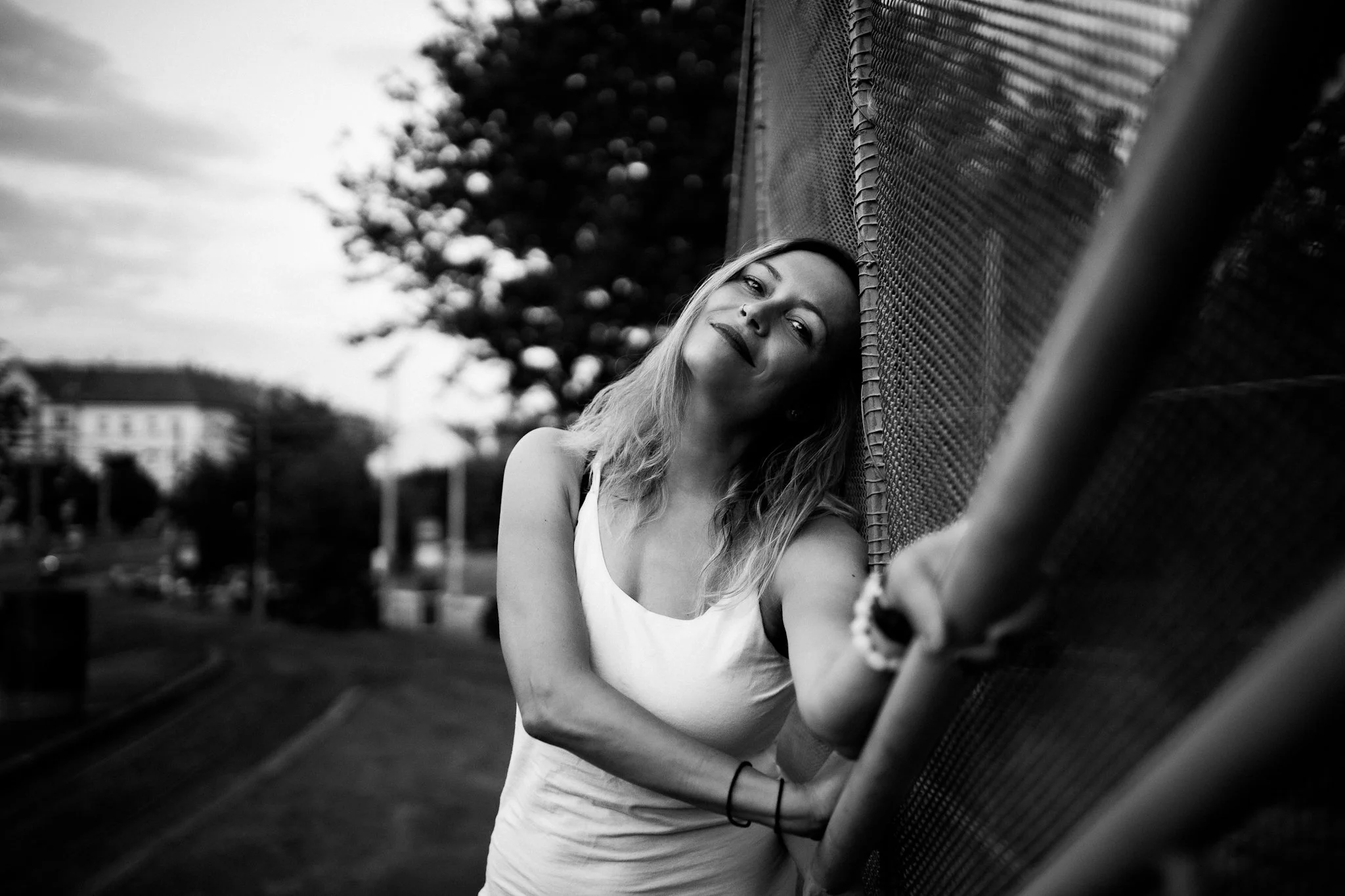 A woman with long wavy hair is leaning against a fence outdoors, looking at the camera with a slight smile. The photo is in black and white, with trees and buildings in the background.