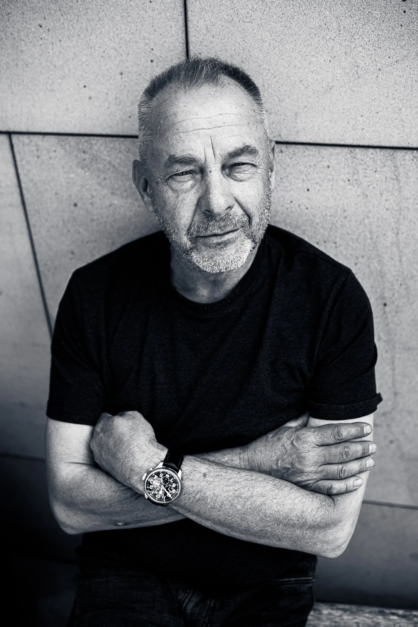 Black and white photo of an older man with short hair and a beard, wearing a wristwatch, black t-shirt, sitting against a textured wall, with arms crossed, looking directly at the camera with a slight smile.