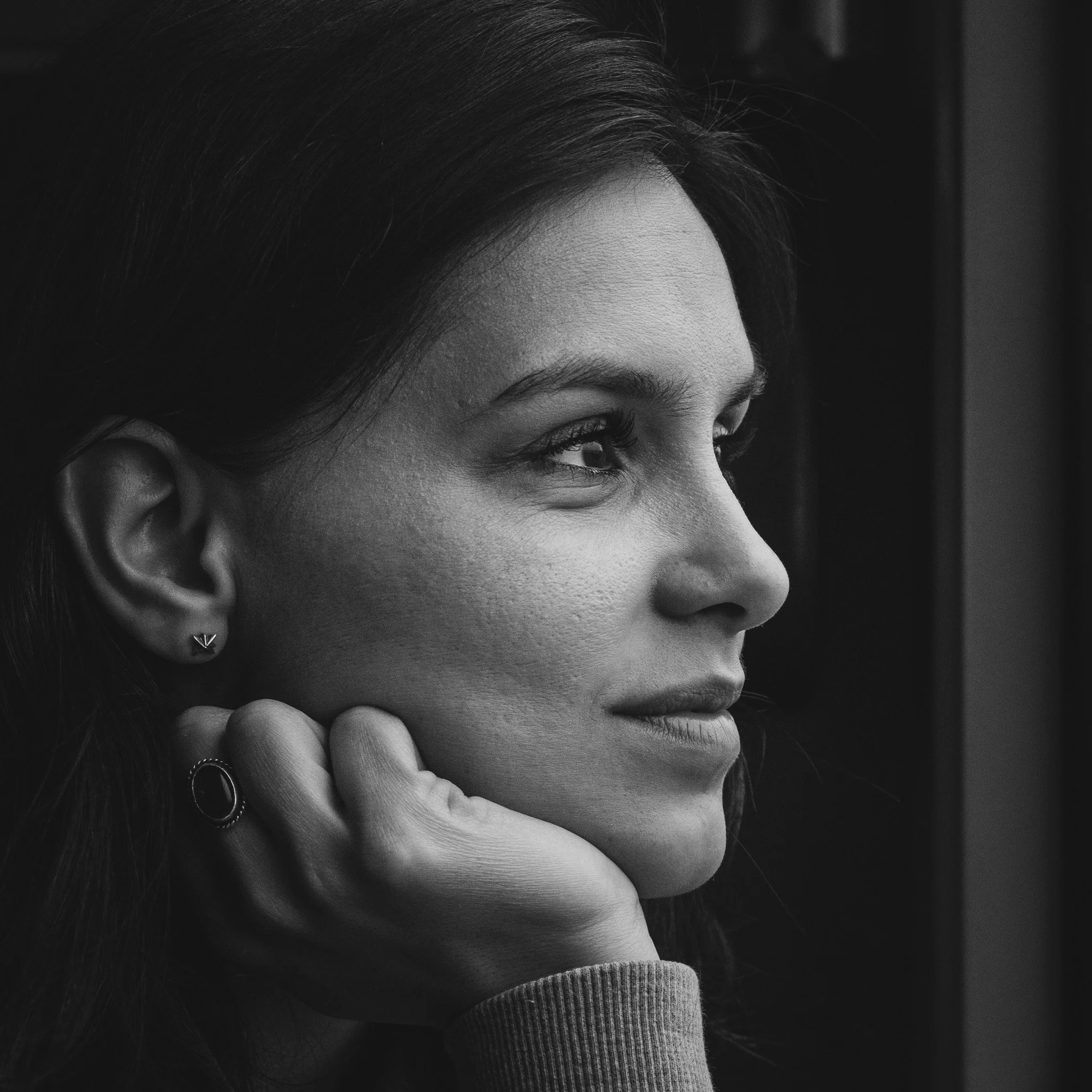 Black and white close-up photo of a woman with dark hair, looking thoughtful, resting her chin on her hand, wearing earrings and rings.