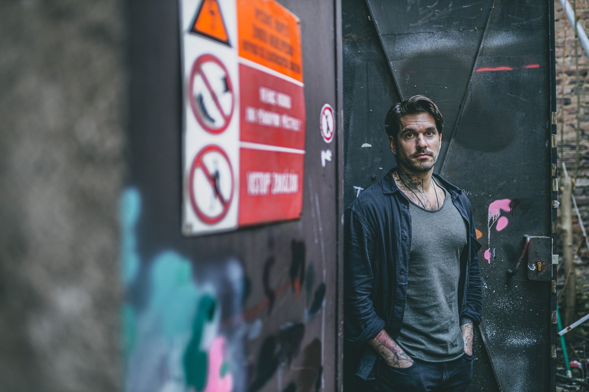 A man with dark hair, tattoos on his neck and arms, wearing a gray shirt and black open shirt, stands against a dark metal gate with graffiti and peeling paint, with warning and prohibition signs on the left side of the image.