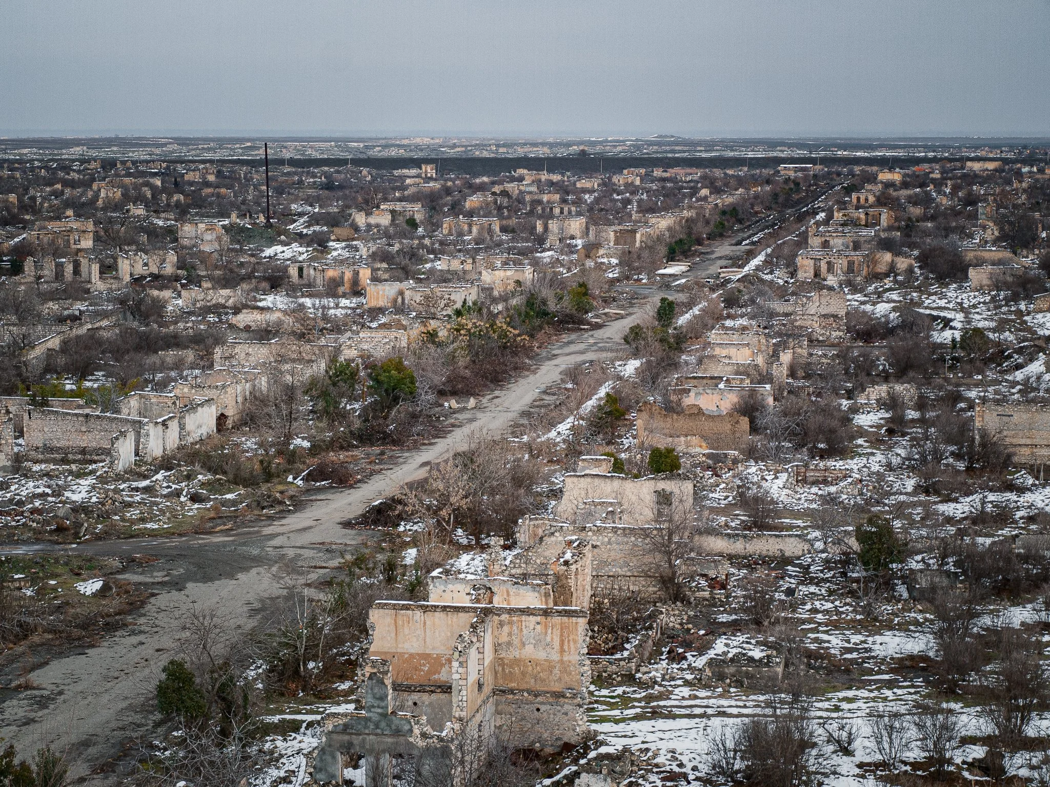 A wide aerial view of a snow-covered, abandoned town with ruined buildings and empty streets stretching into the distance under a cloudy sky.