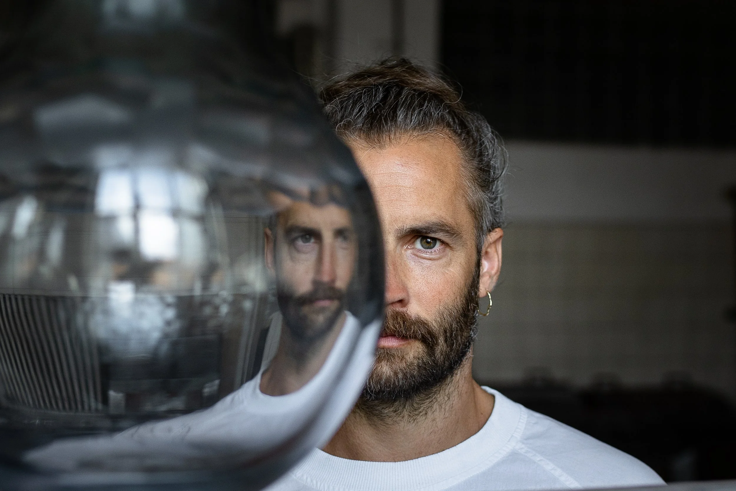 A man with a beard, wearing a white shirt, looking at the camera with a mirror reflecting his face.