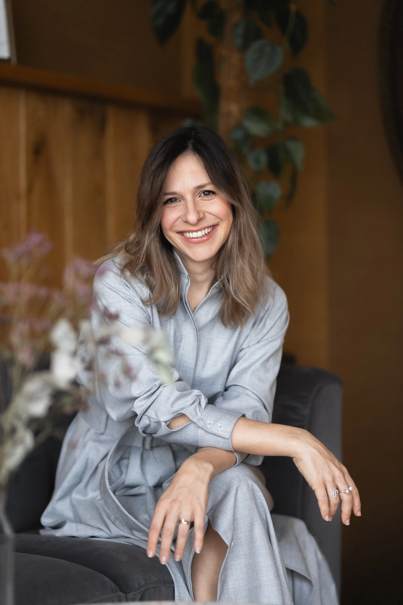 A woman with long, wavy brown hair, smiling, and sitting on a black sofa in a cozy room with wooden walls and green plants.