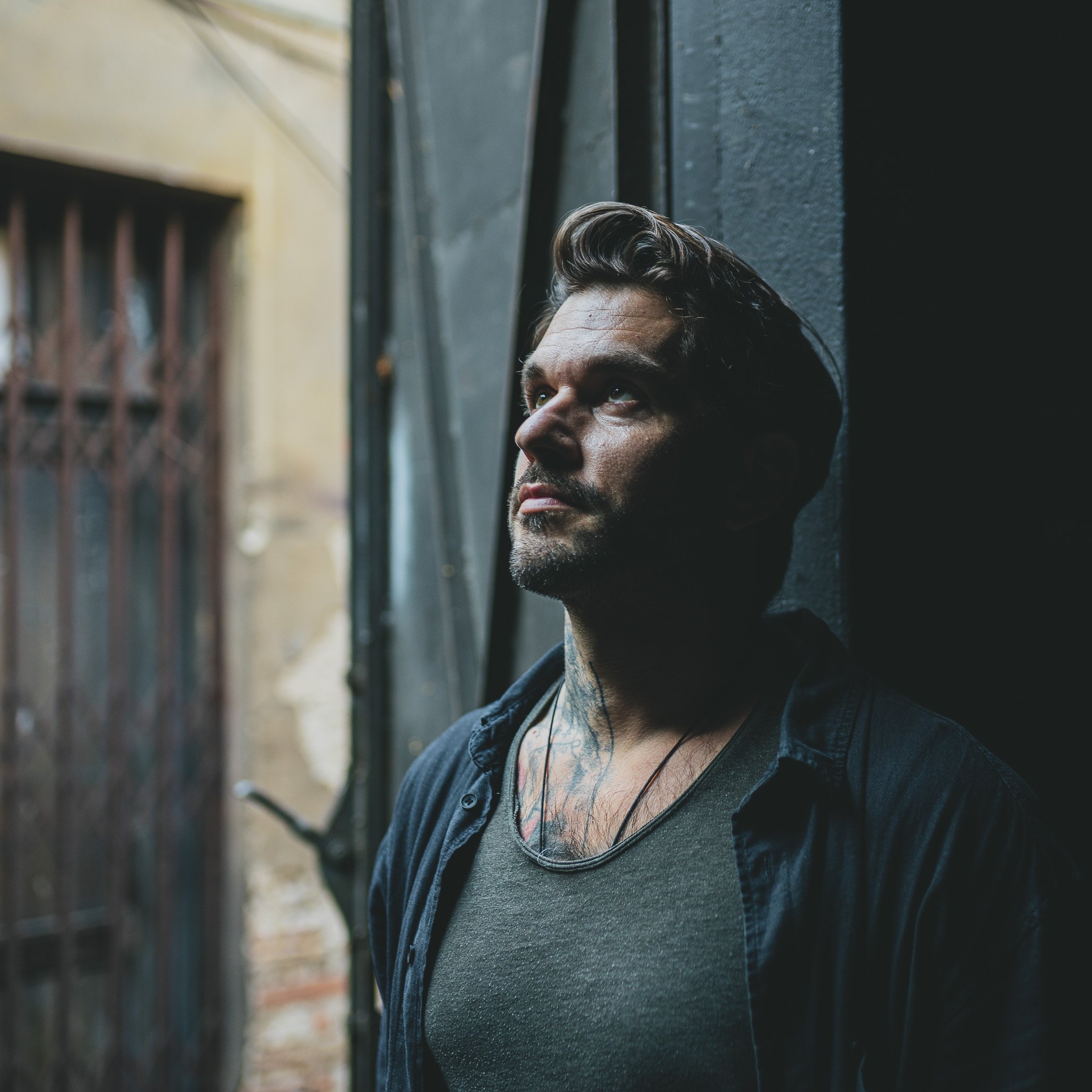 A man with dark hair and tattoos gazes upward while leaning against a dark-colored wall in an urban setting.