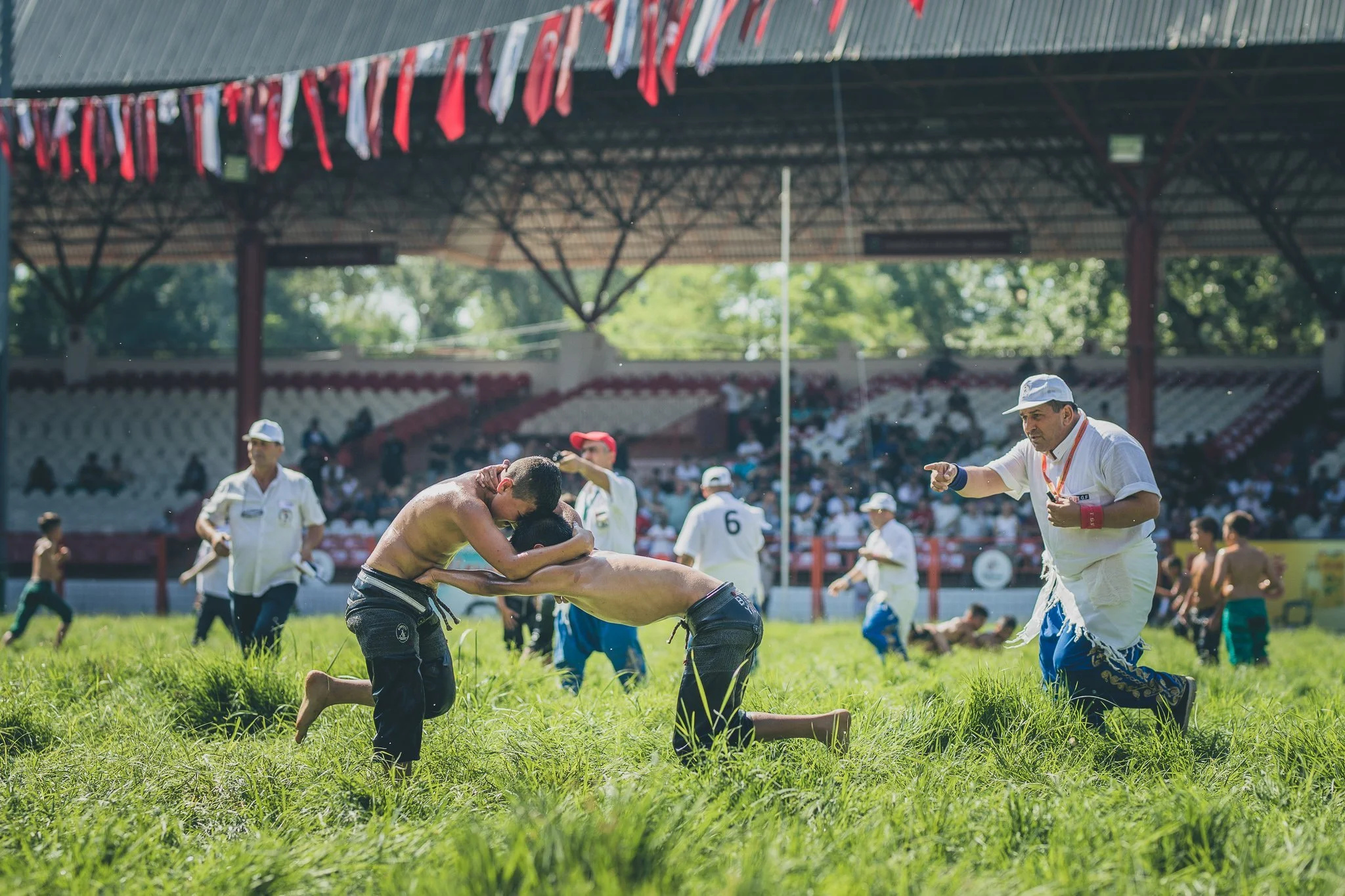 Two men wrestling on a grassy field during a traditional sports event, surrounded by several spectators and officials under a decorated pavilion with red and white banners.