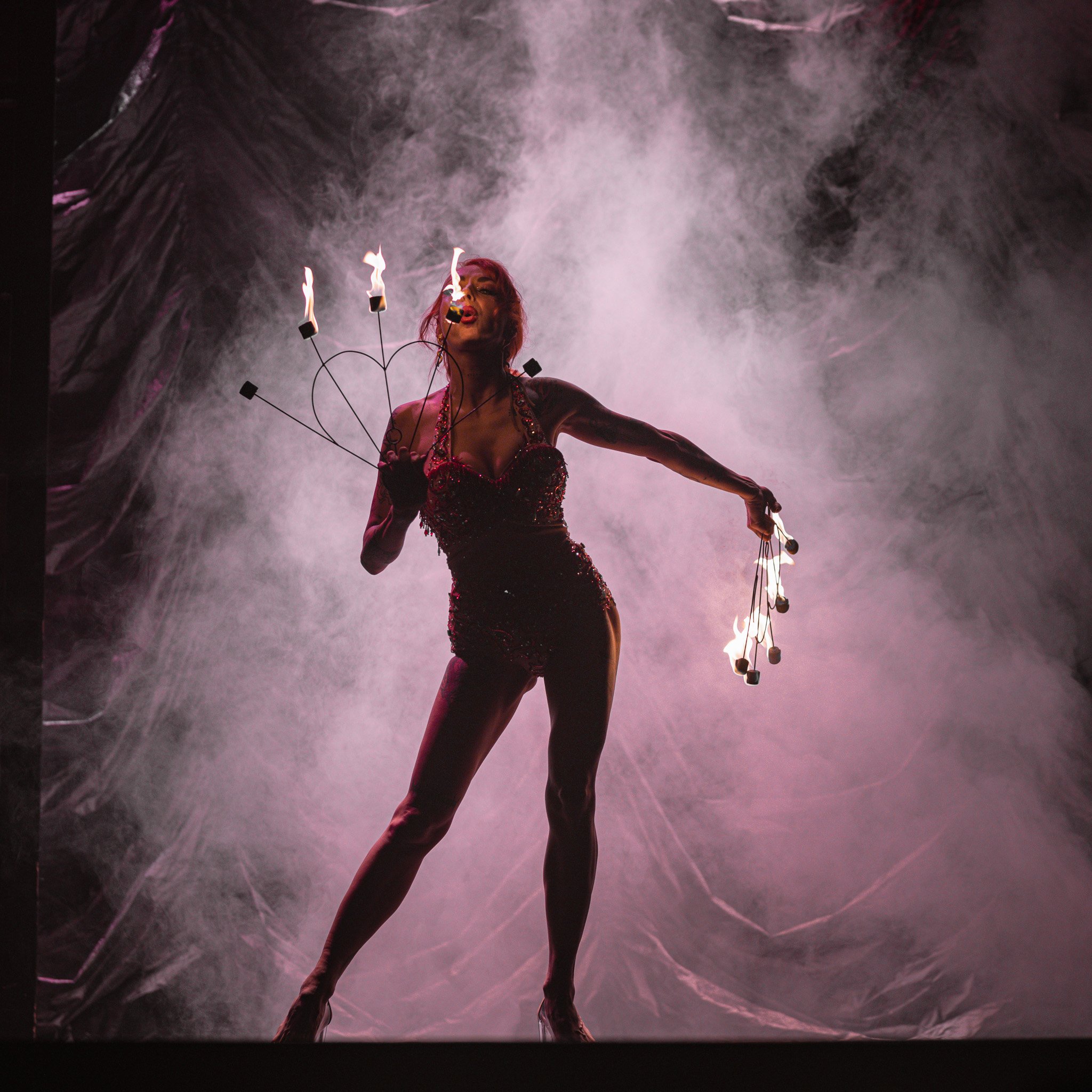 Performer in a dark, glittering costume holds flaming torches in both hands on stage with smoky background