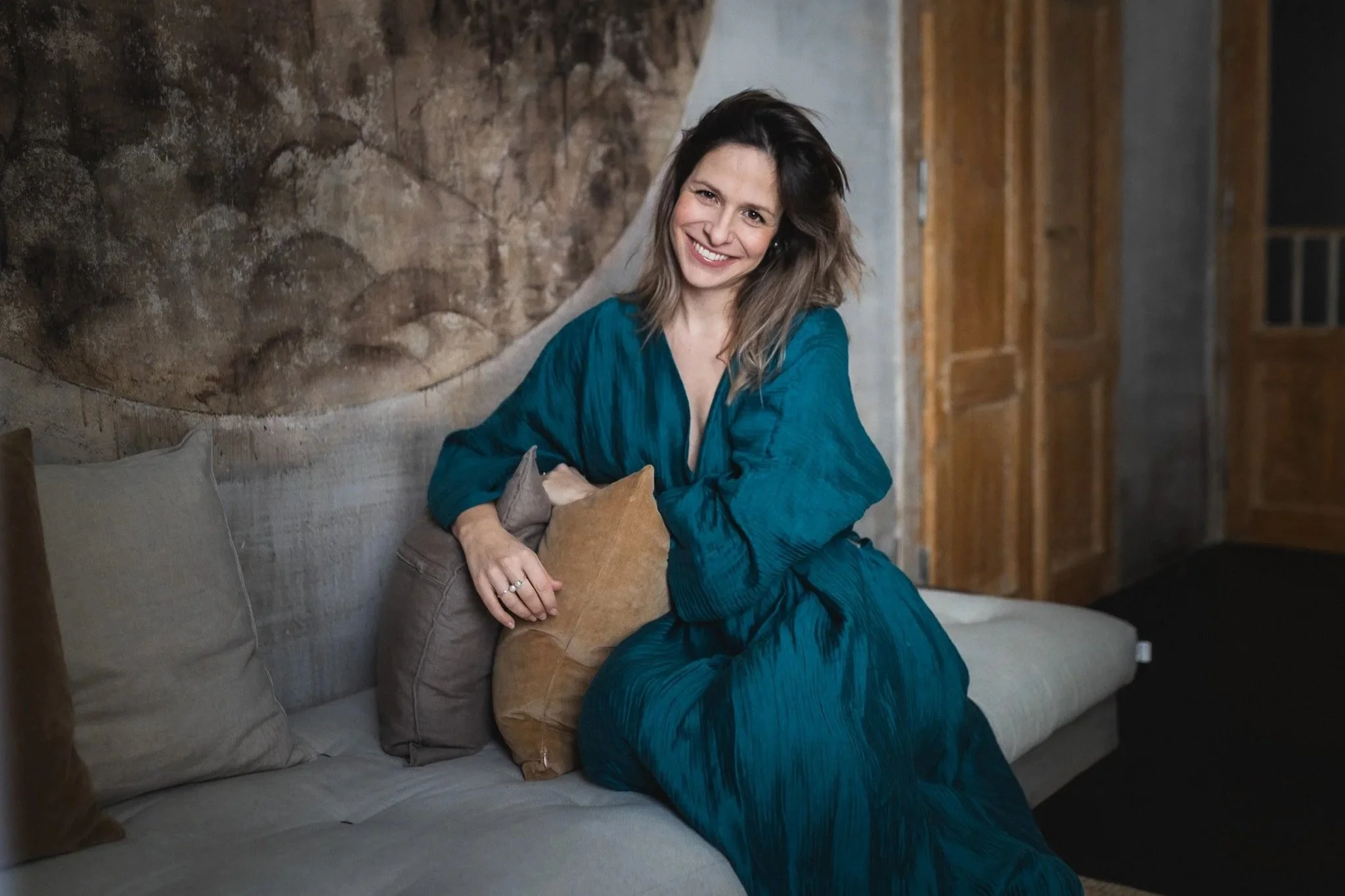 A woman in a teal dress sitting on a beige couch, smiling, with a dark, artistic wall behind her and wooden doors in the background.