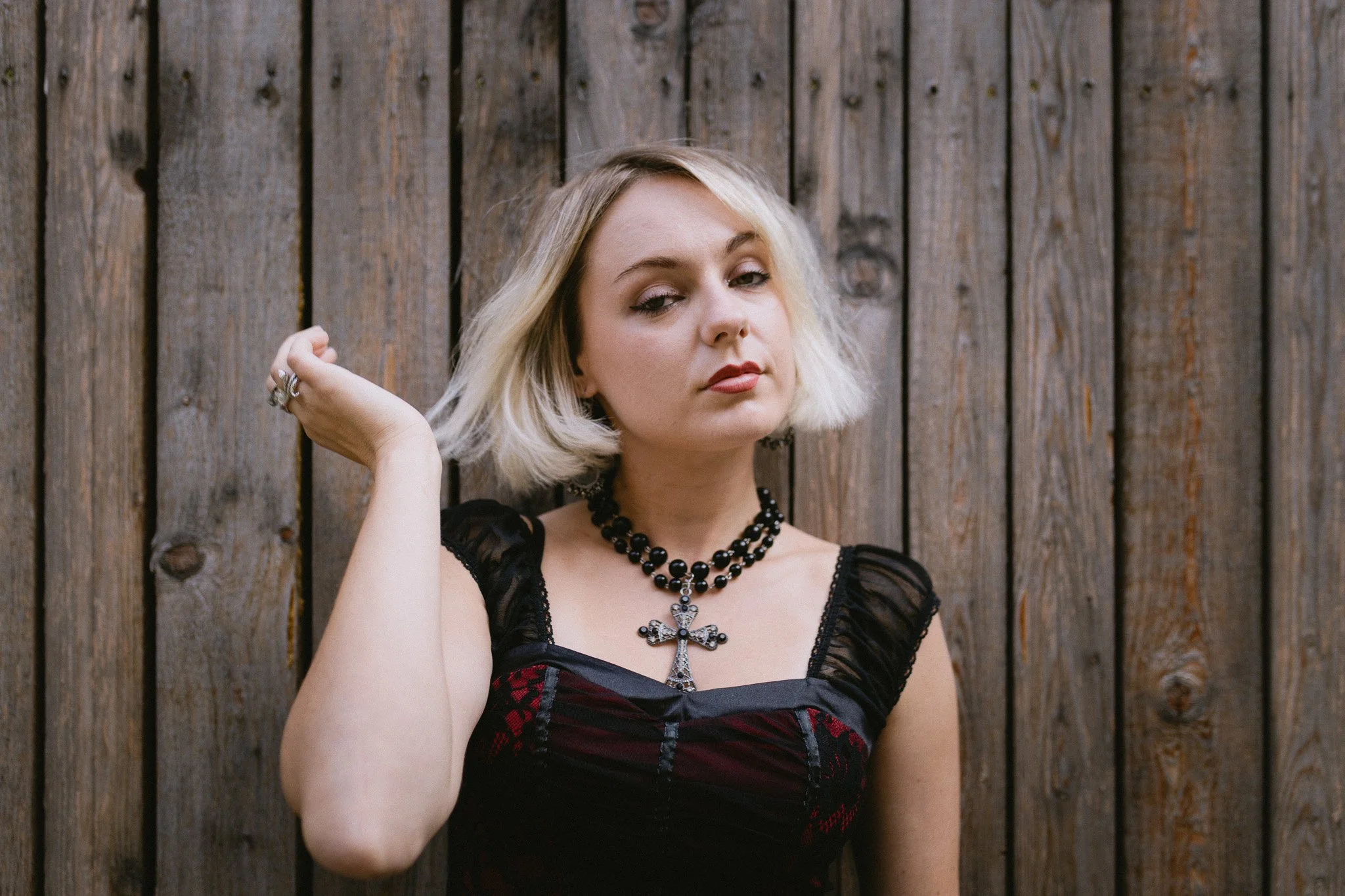 A woman with blonde hair and tattoos, wearing a black dress with lace details and a large cross necklace, standing in front of a wooden fence, looking at the camera with a confident expression.