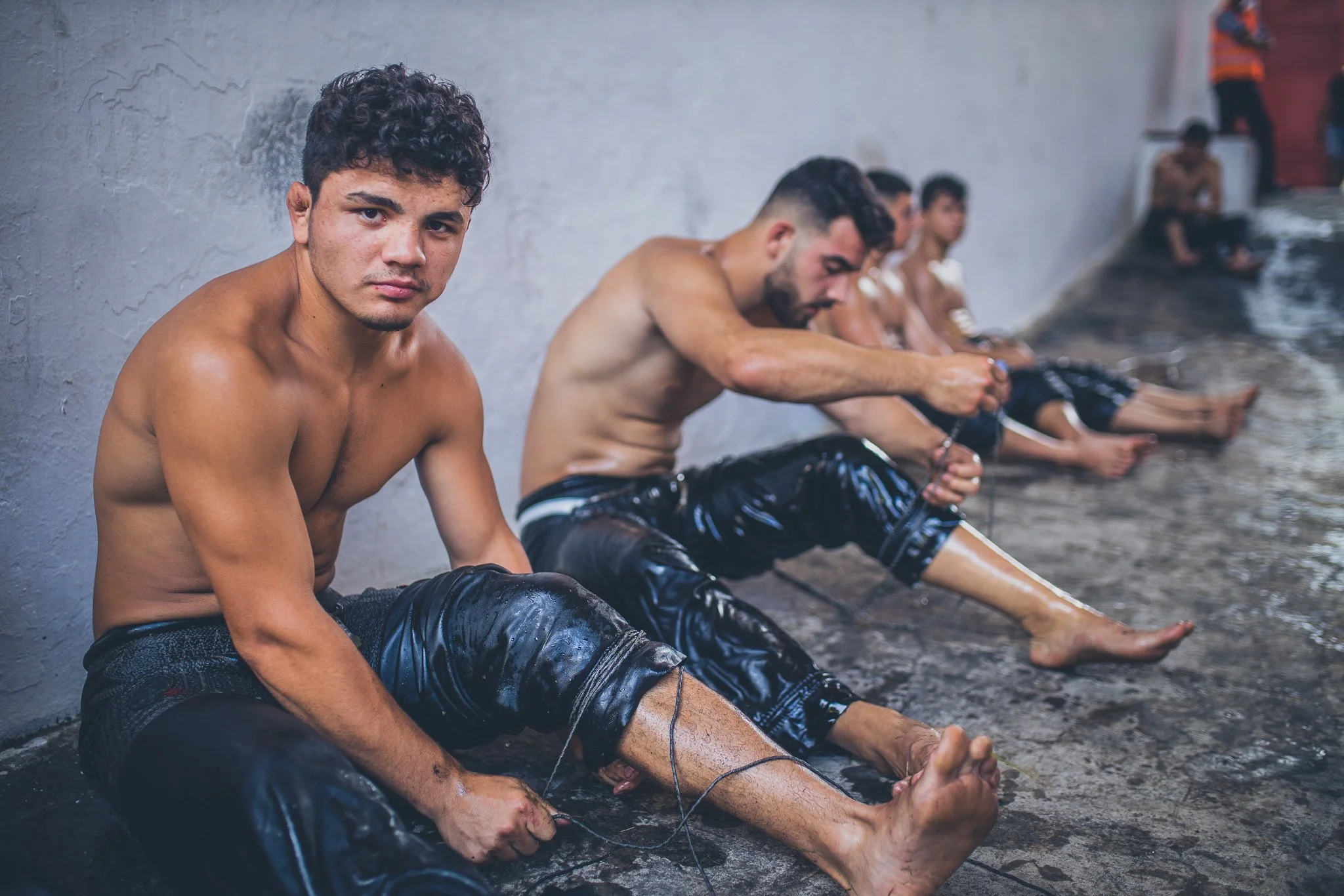 Young group of men sitting on the ground by a pool, wearing black pants, with some of them in the water, tying white string around their ankles.