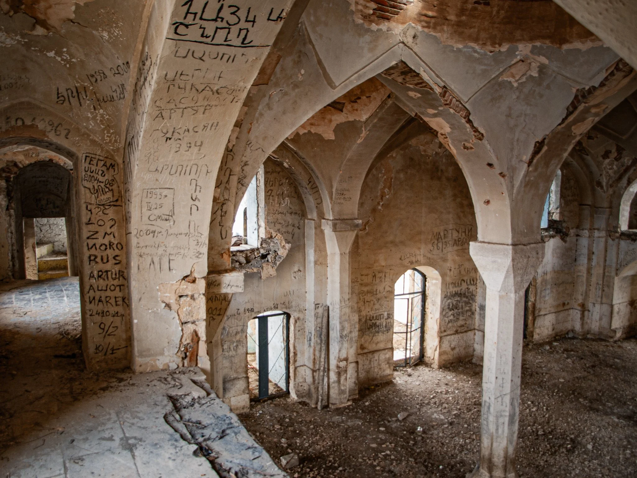 Interior of an old stone mosque with arches covered in graffiti, some windows with metal bars, and a dirt floor.