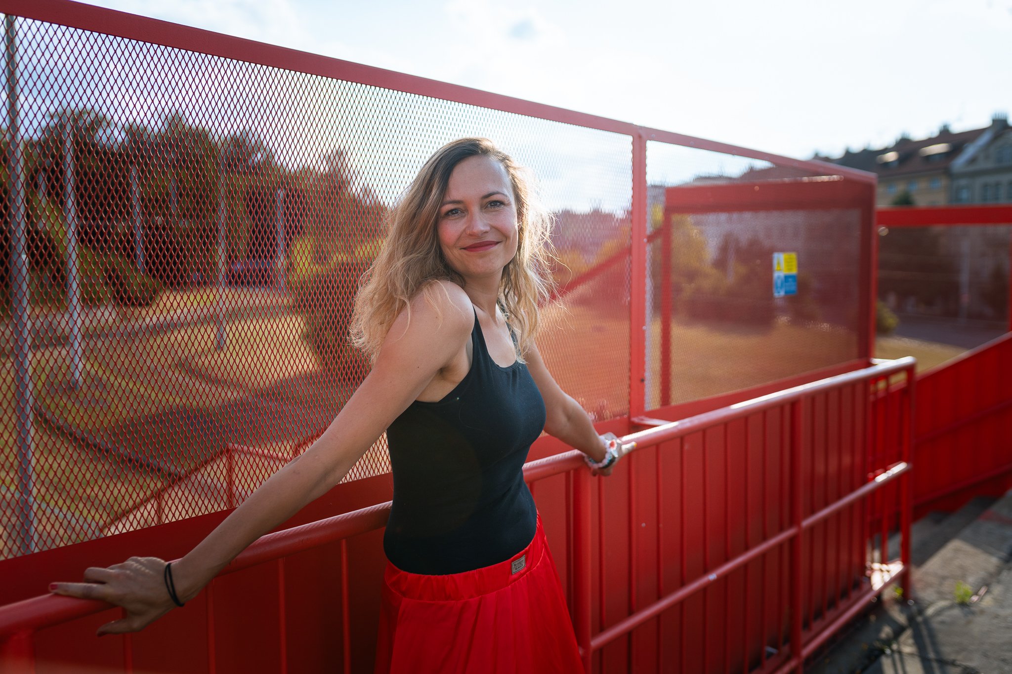 A woman with blonde hair and a black tank top leans on a red railing outside, smiling at the camera with a background of trees and houses.