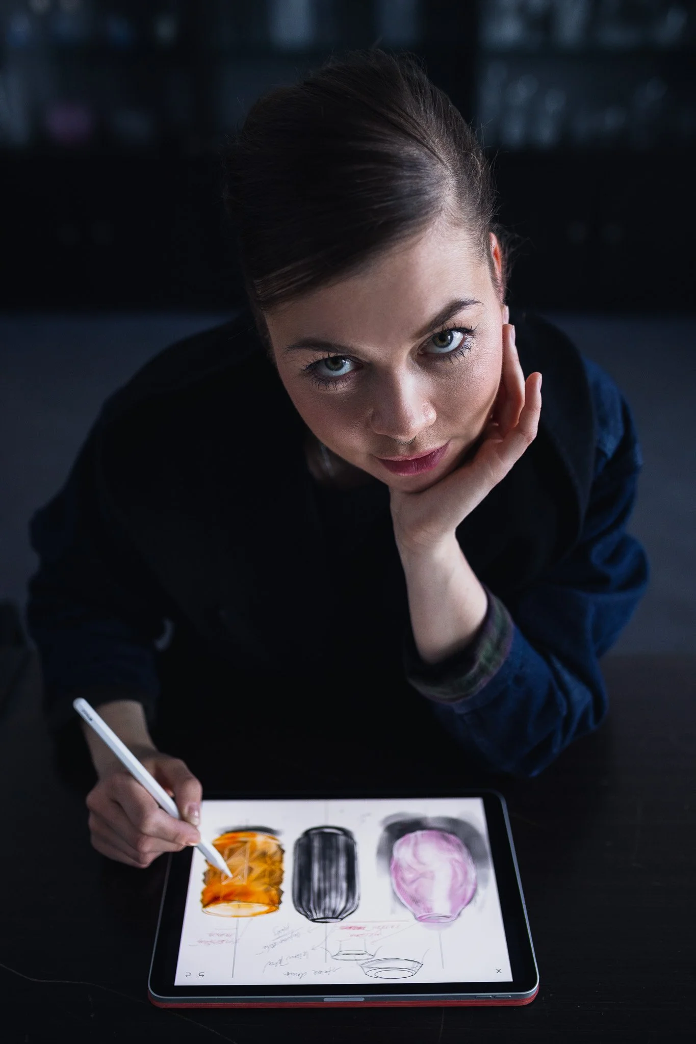 A woman with brown hair and blue eyes drawing colorful glasses designs on a tablet with a stylus, resting her head on her hand, in a dark environment.