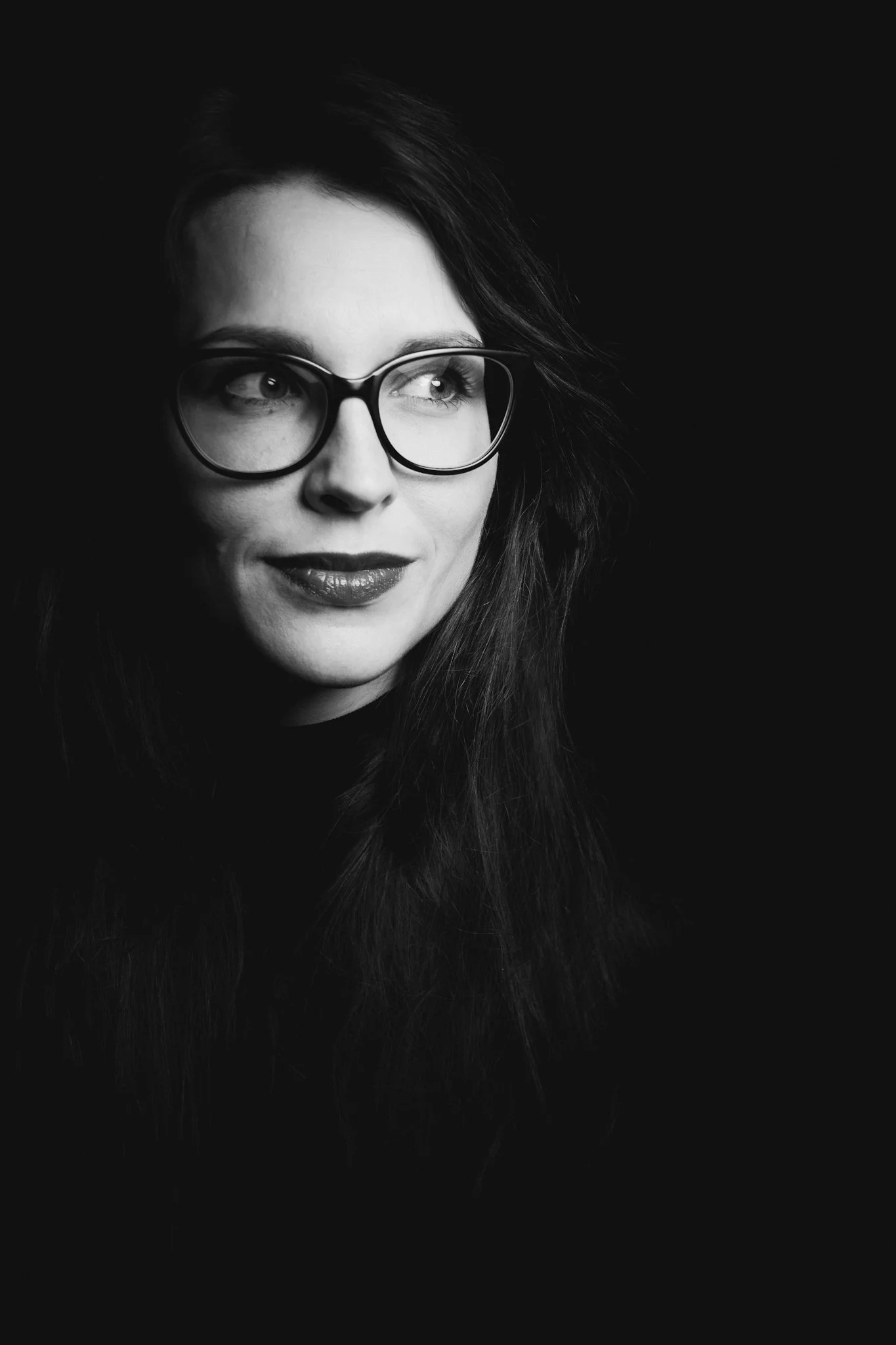 Black and white portrait of a woman wearing glasses, with long hair, looking to the side, against a dark background.