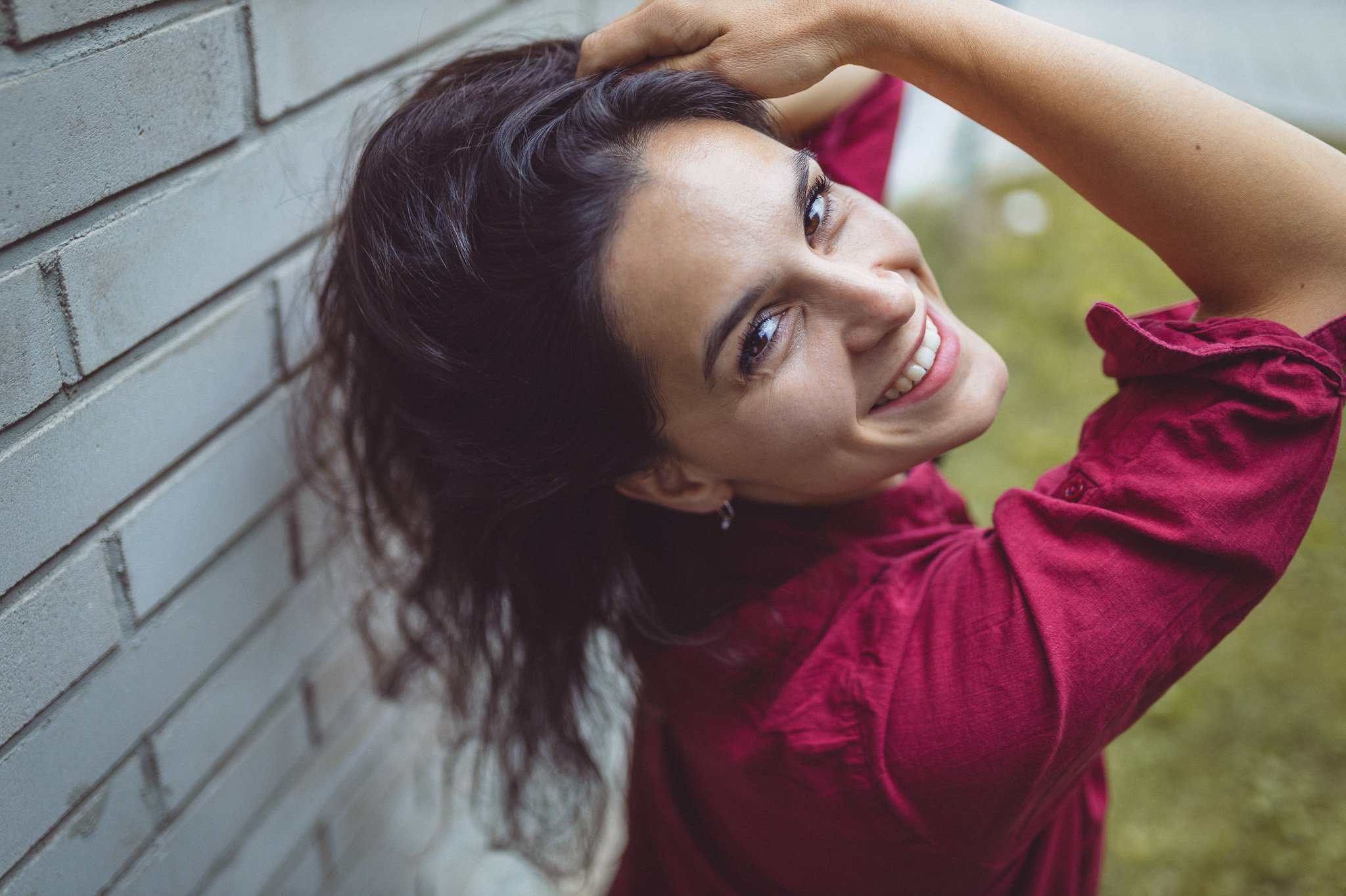 A woman with dark hair and a red shirt leaning against a gray brick wall, smiling and looking up.