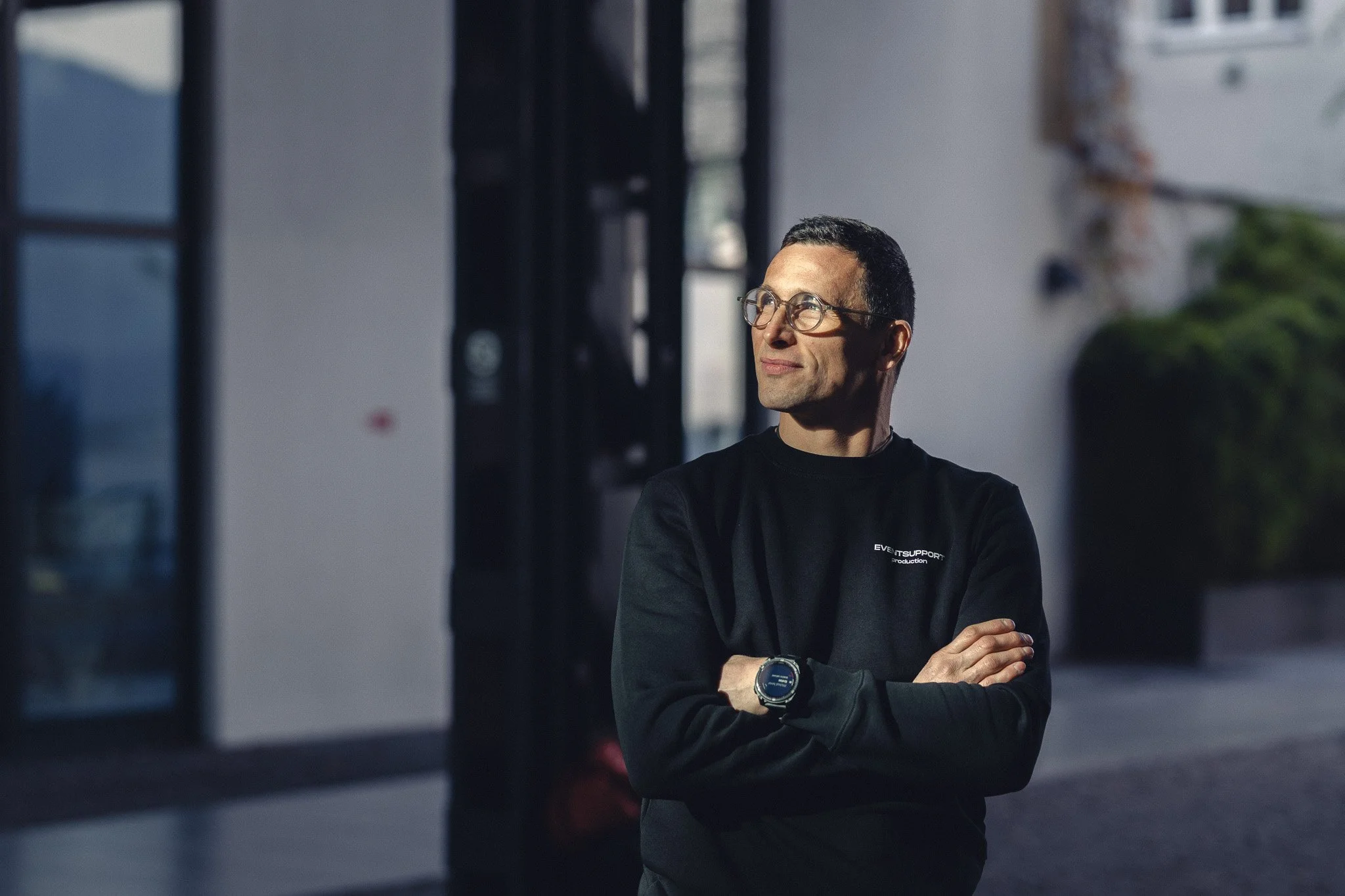 A man with glasses and a black sweatshirt with white text stands outdoors with arms crossed, looking to the side, illuminated by sunlight.