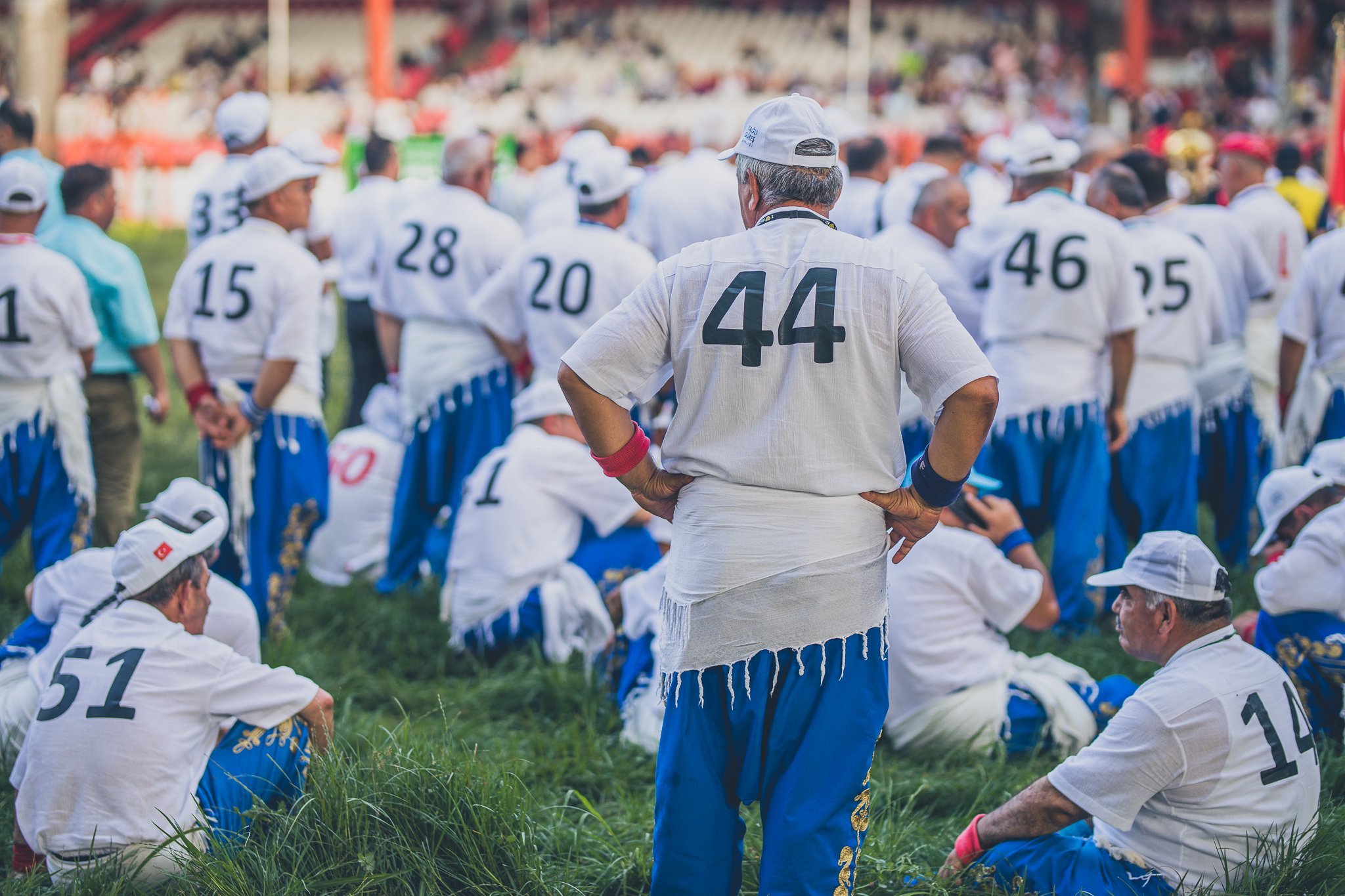 People wearing white shirts and blue pants gathered outdoors, some sitting on grass and others standing, at a festival or gathering.