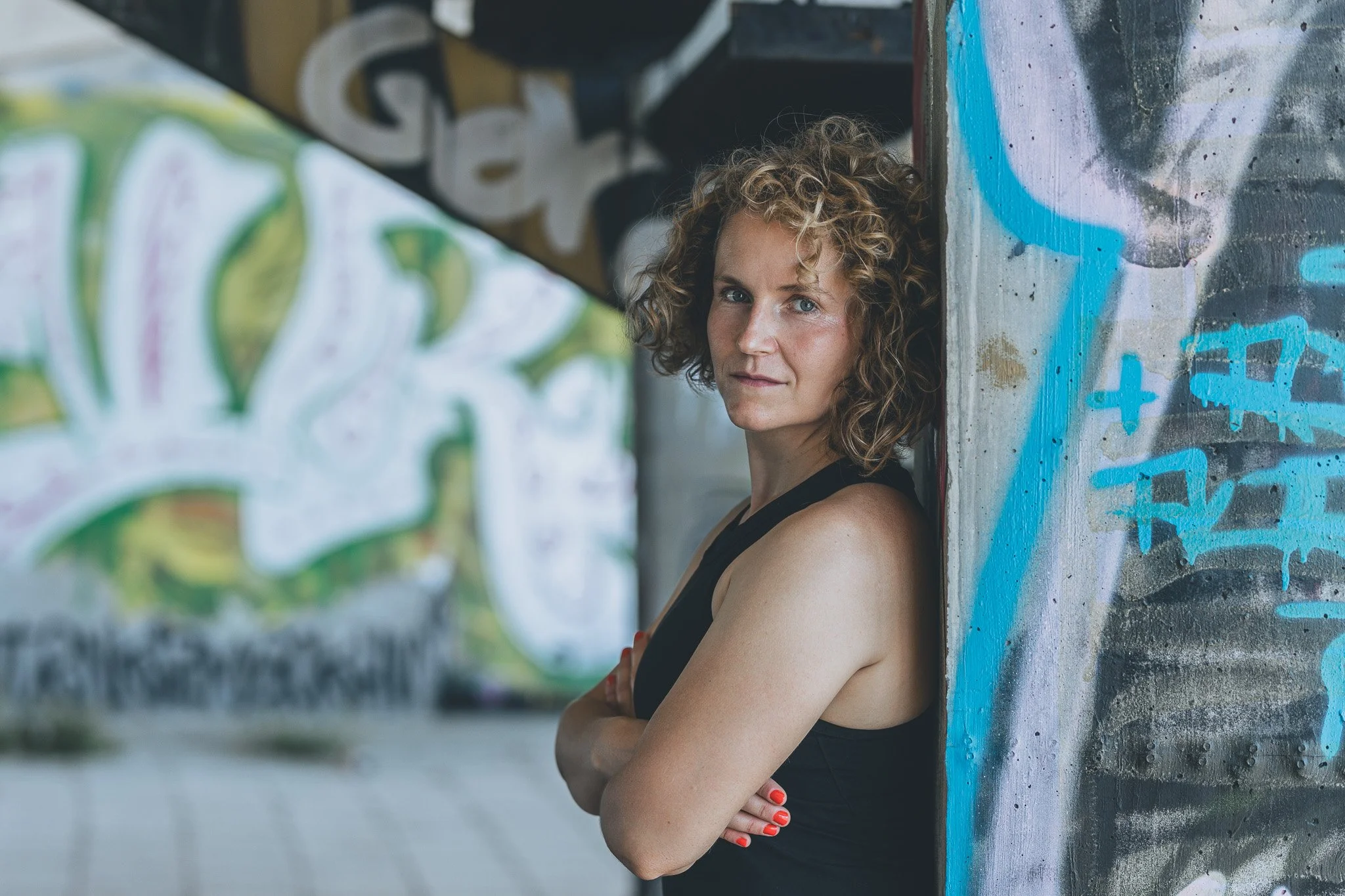 A woman with curly hair leaning against a graffiti-covered wall, looking at the camera.