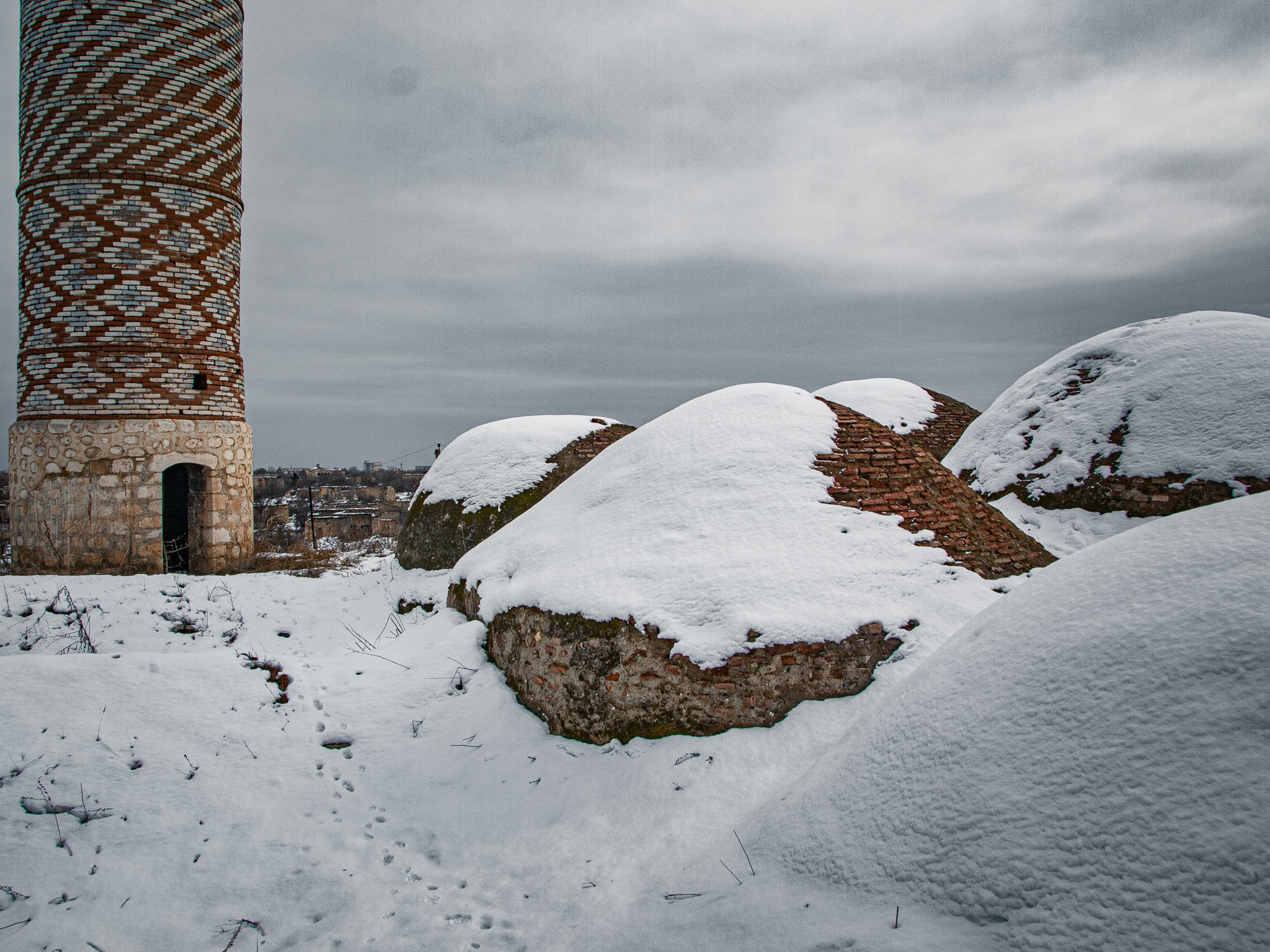 Snow-covered ruins with a tall brick tower on the left and large, rounded brick structures on the ground, under a cloudy sky.
