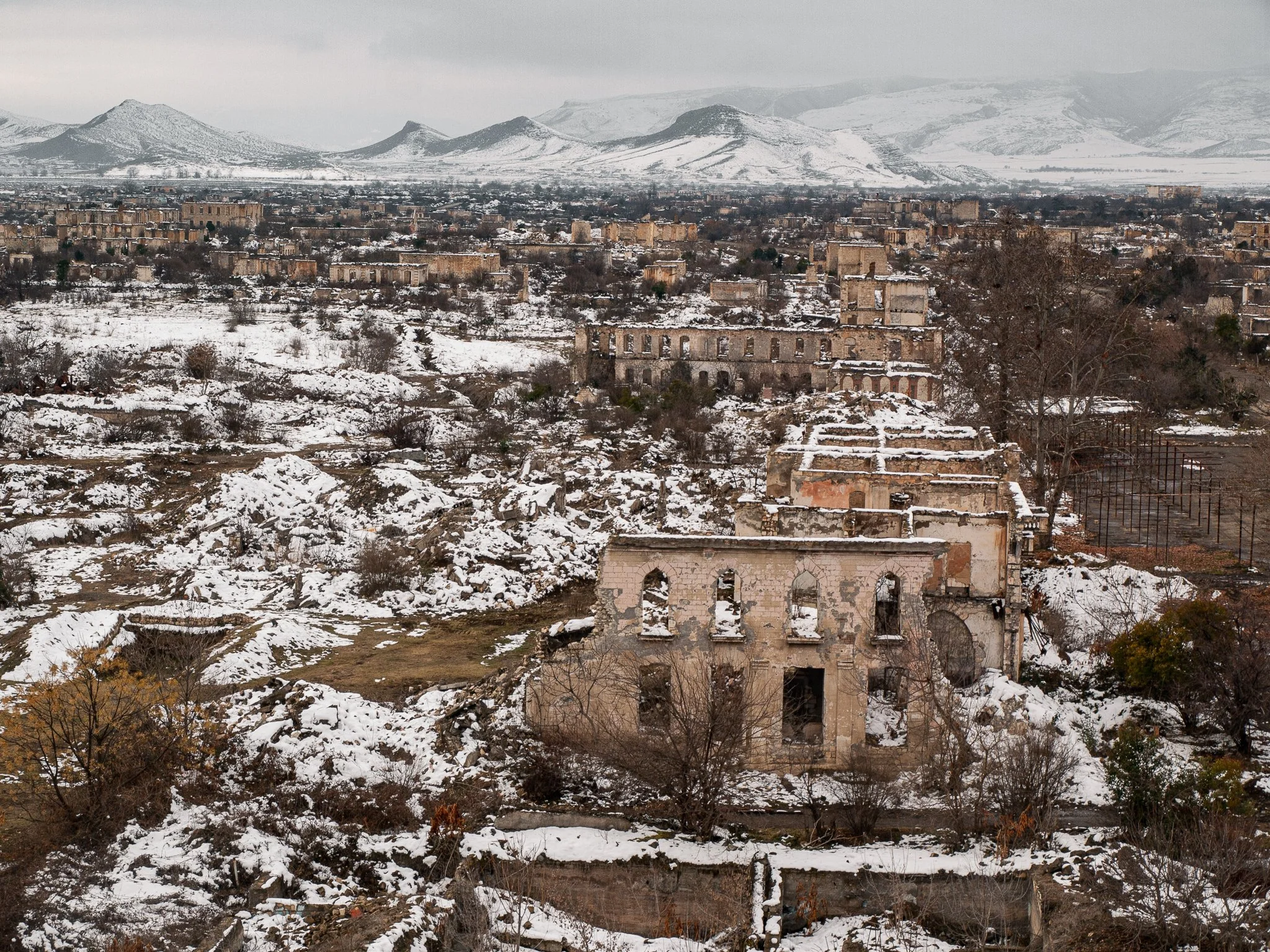 Snow-covered ruins of an abandoned building in a desolate landscape, with a city and snow-capped mountains in the background.