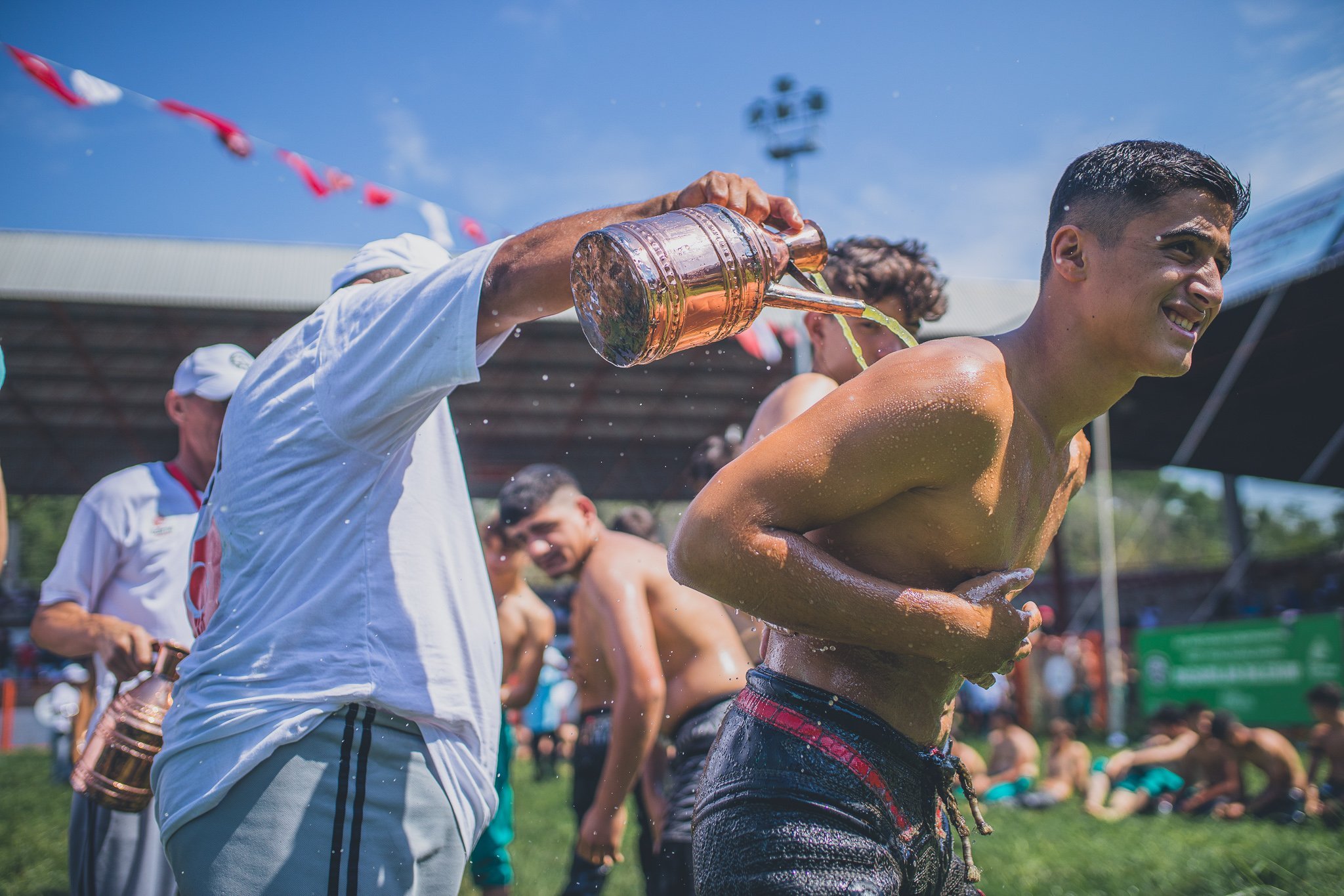 Young man running outdoors, shirtless, with water being poured on him, others in the background, sunny day, celebrating or cooling off.