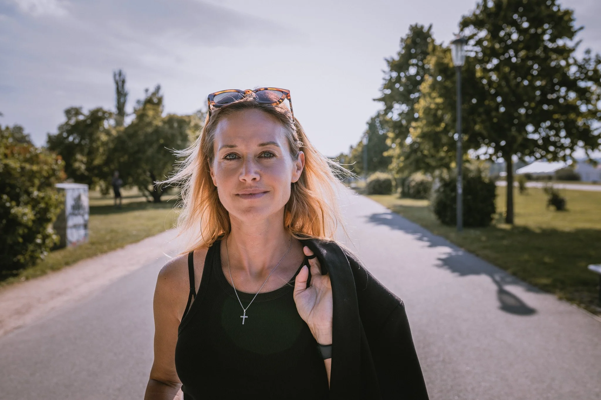 A woman with blonde hair, wearing sunglasses on her head, a black tank top, and a silver cross necklace, standing outdoors on a sunny day in a park, holding a black jacket over her shoulder.