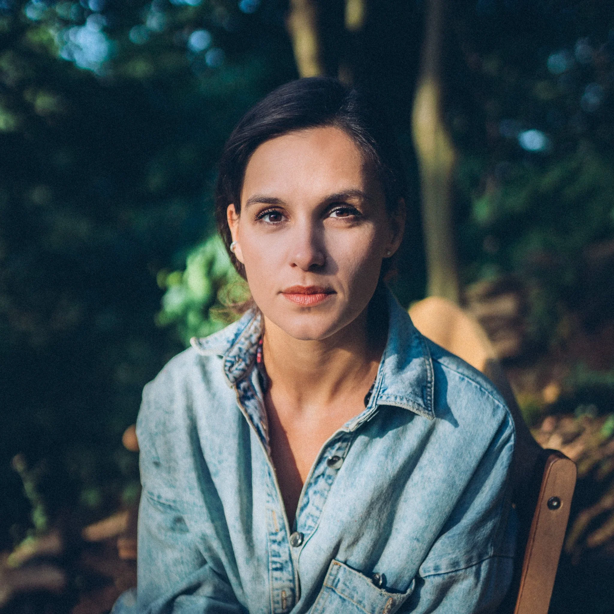 A woman with short dark hair and light skin sitting outdoors in a natural setting, wearing a denim shirt.