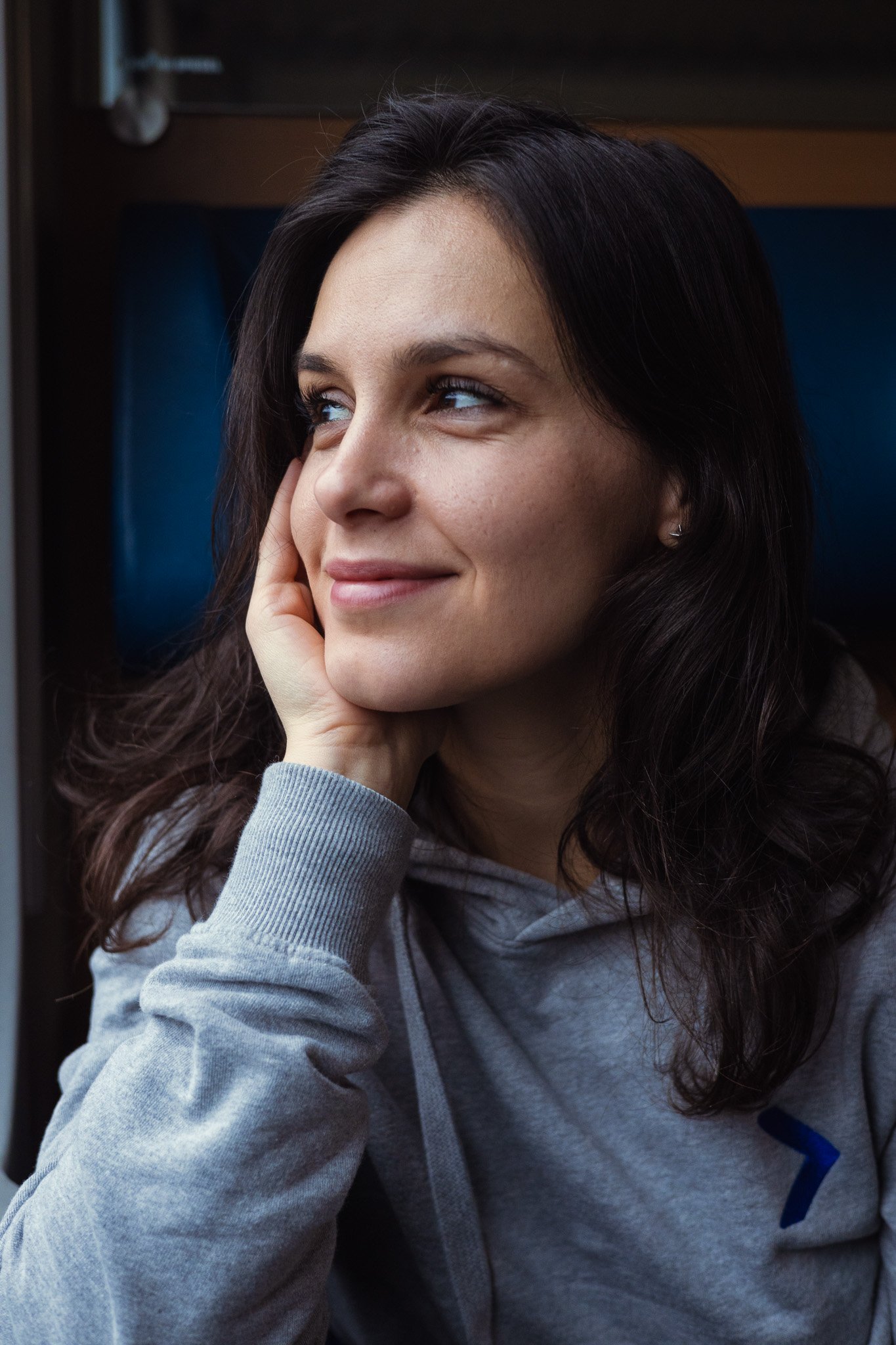 A young woman with dark hair and blue eyes smiling, resting her face on her hand, wearing a gray hoodie.
