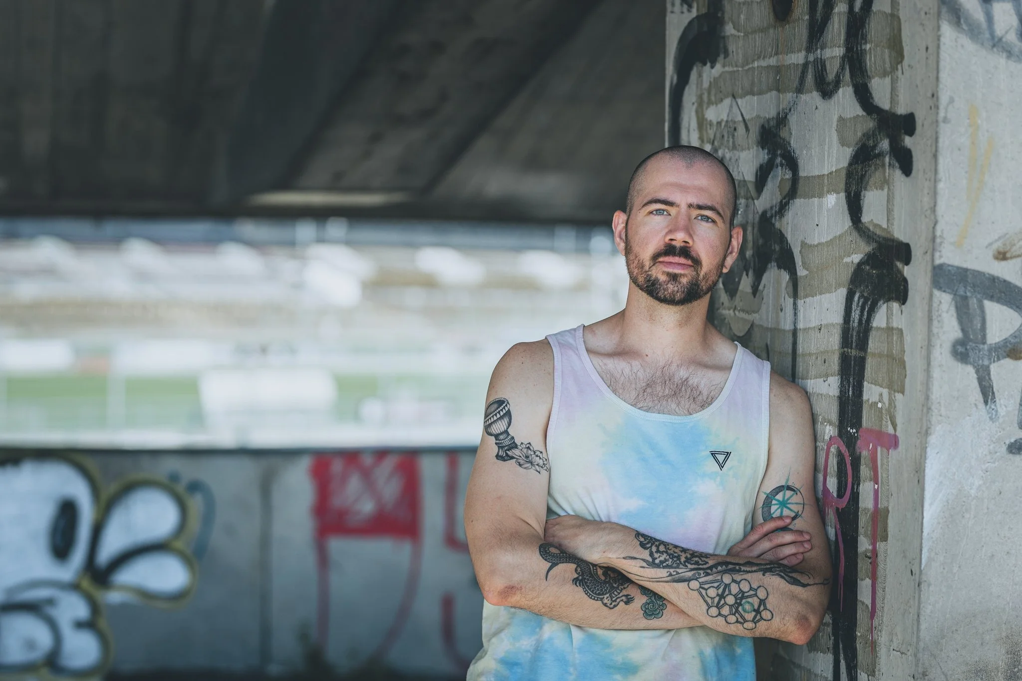 A man with a beard and tattoos on his arms, wearing a tie-dye tank top, standing with arms crossed against a graffiti-covered concrete wall under a bridge or overpass.