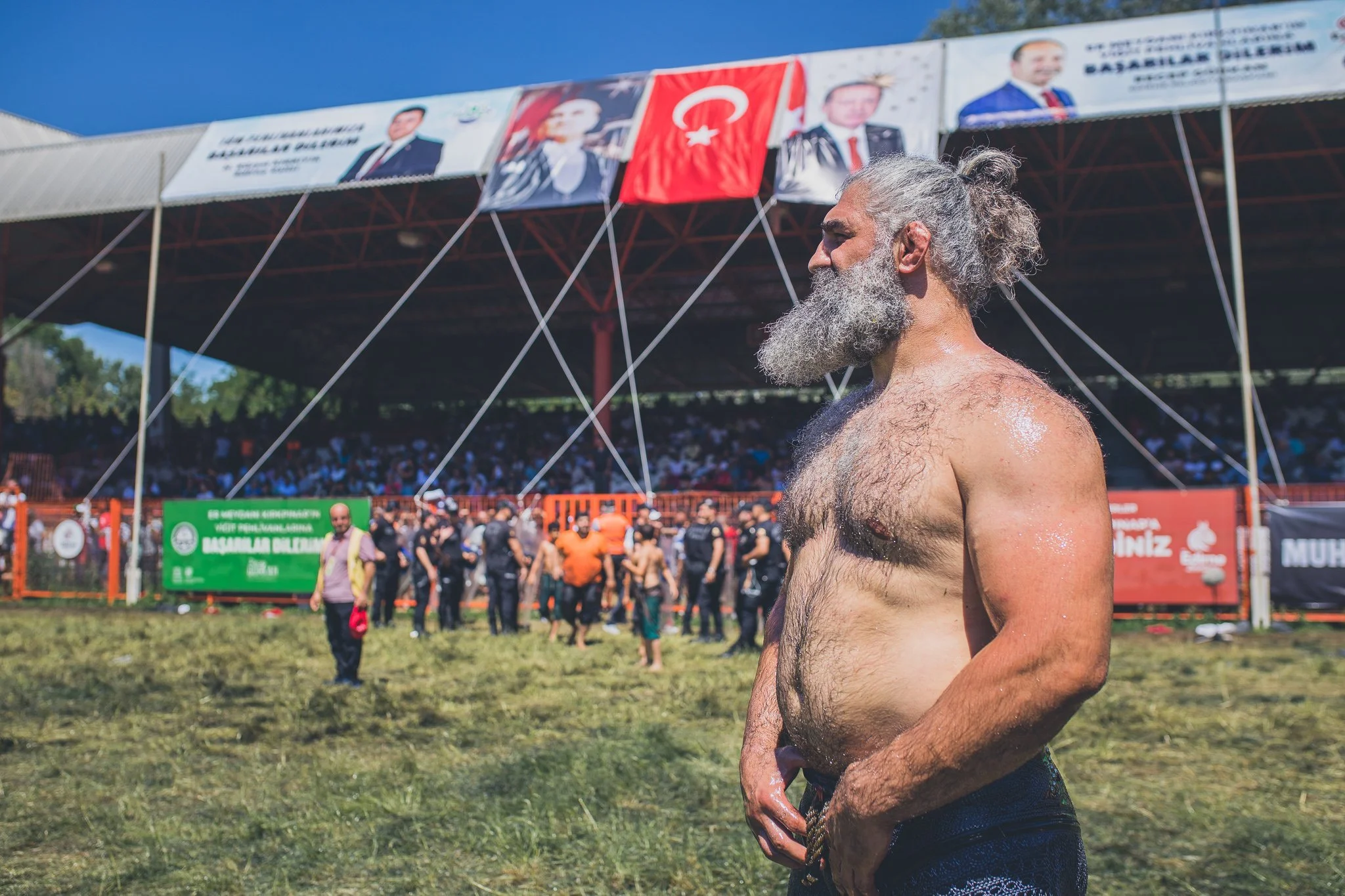 An older, shirtless man with a long gray beard and hair tied in a bun stands outdoors on a grassy field, looking to the side. Behind him, there is a crowd of people and a large stage decorated with banners and a Turkish flag under a partly cloudy sky