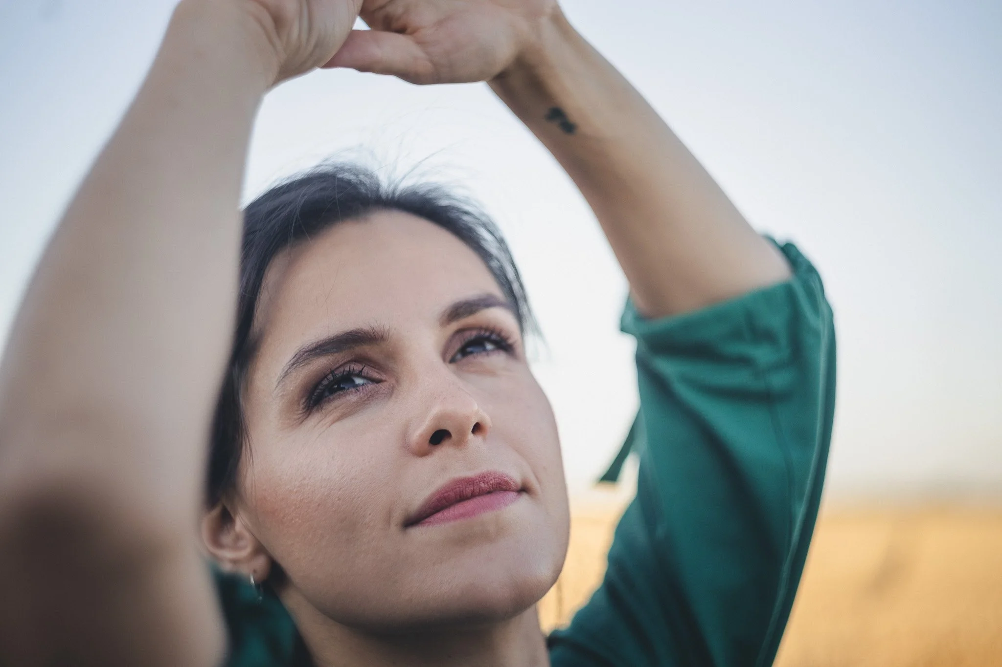 A woman with dark hair and light makeup is raising her arms above her head, looking up into the sky with a peaceful expression, outdoors during daytime.