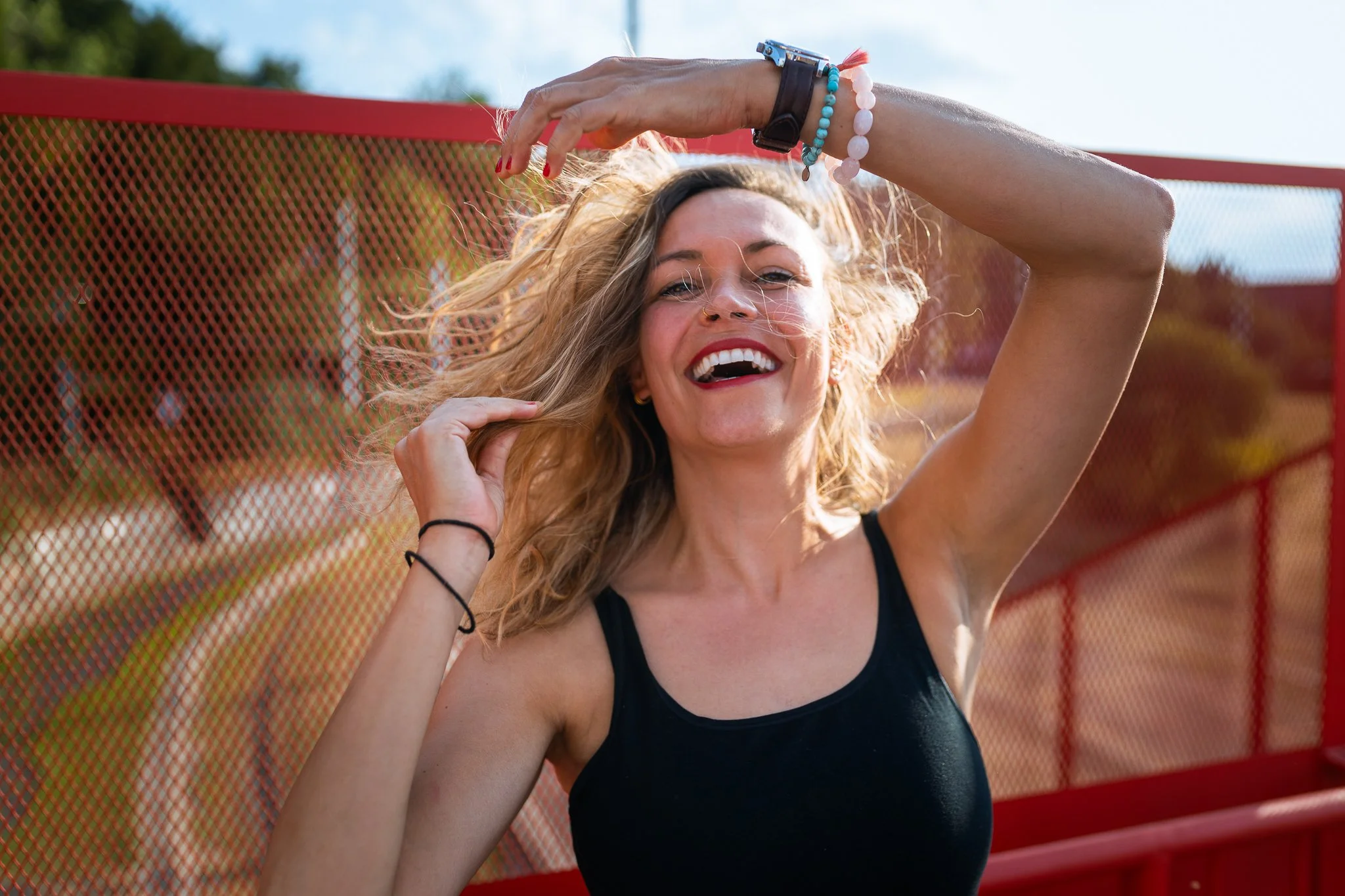 A young woman with blonde curly hair, smiling and posing outdoors near a red fence, wearing a black tank top, with some bracelets on her wrist.