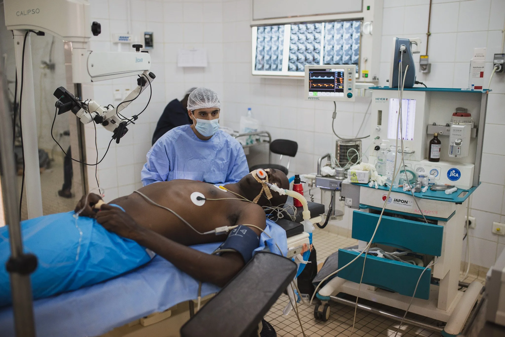 A medical team monitors a patient in an intensive care unit. The patient, a man, is lying on a hospital bed connected to various medical devices and tubes, with a mask on his face. A healthcare worker in scrubs, a mask, and a hairnet is sitting besid