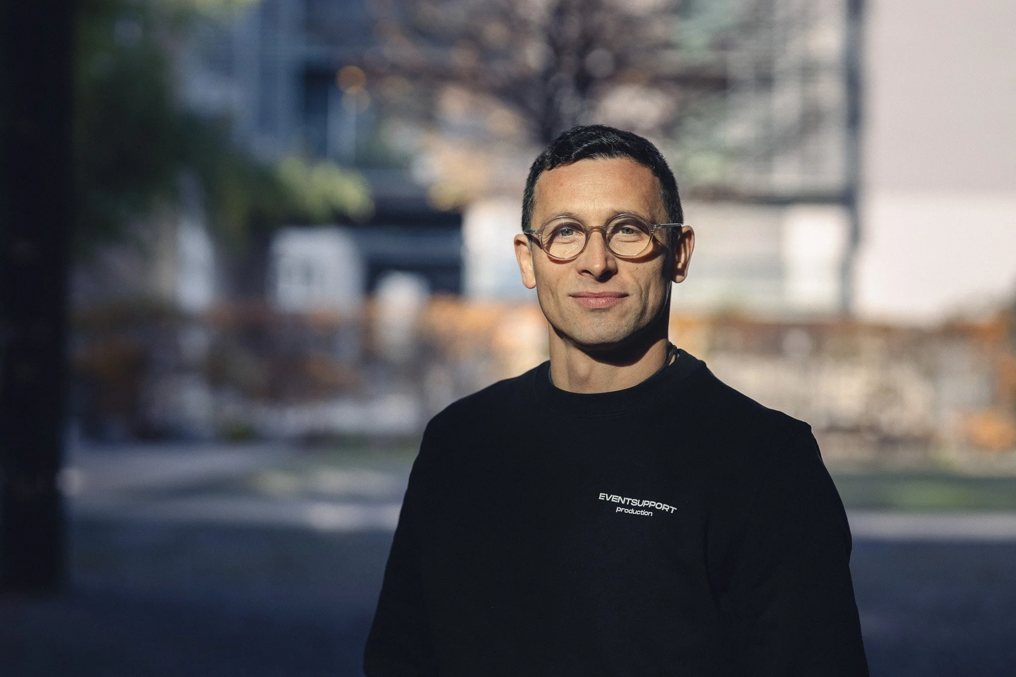 A man with short dark hair, wearing glasses and a black t-shirt with white text that reads 'EVENTSUPPORT production,' standing outdoors during daytime with a blurred background of trees and buildings.