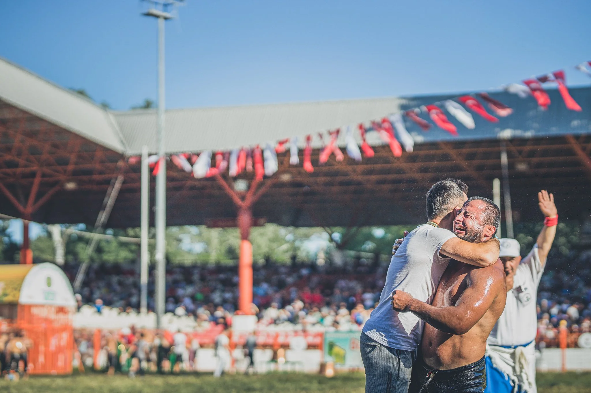 Two men embrace in a celebratory hug at a sports event, with a crowd and a red canopy in the background.