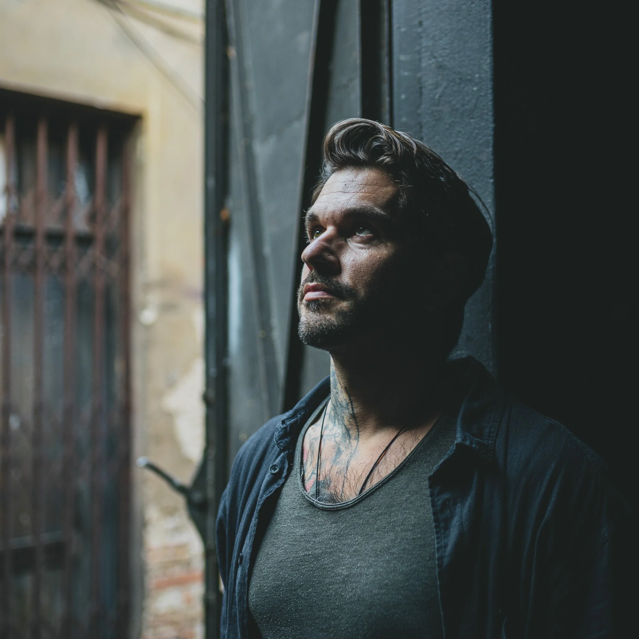 A man with tattooed neck leaning against a dark wall, looking upward with a contemplative expression, near an outdoor gate.
