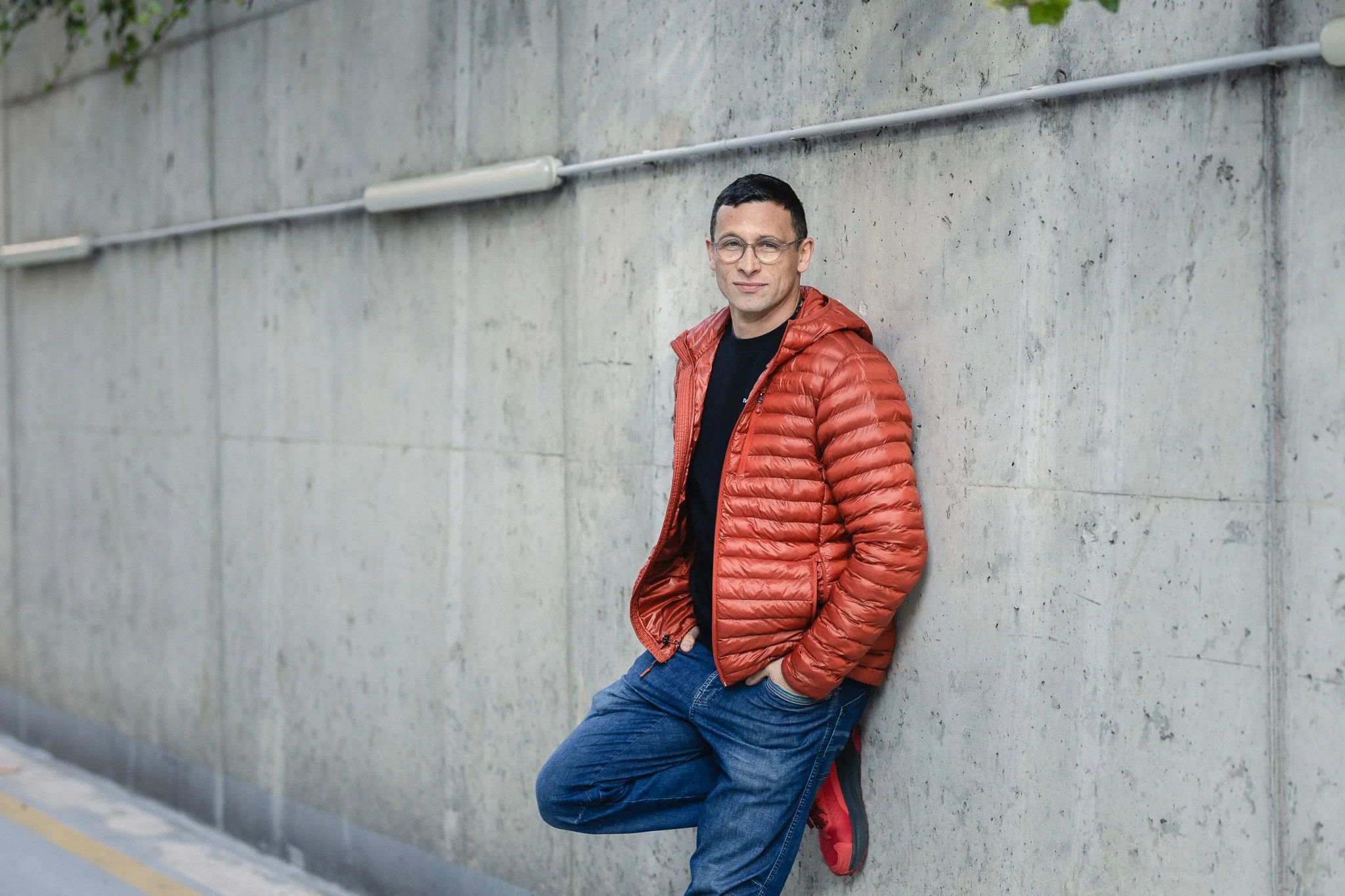 A man leaning against a concrete wall outdoors, wearing glasses, a red puffer jacket, black shirt, and blue jeans, with one foot crossed over the other.