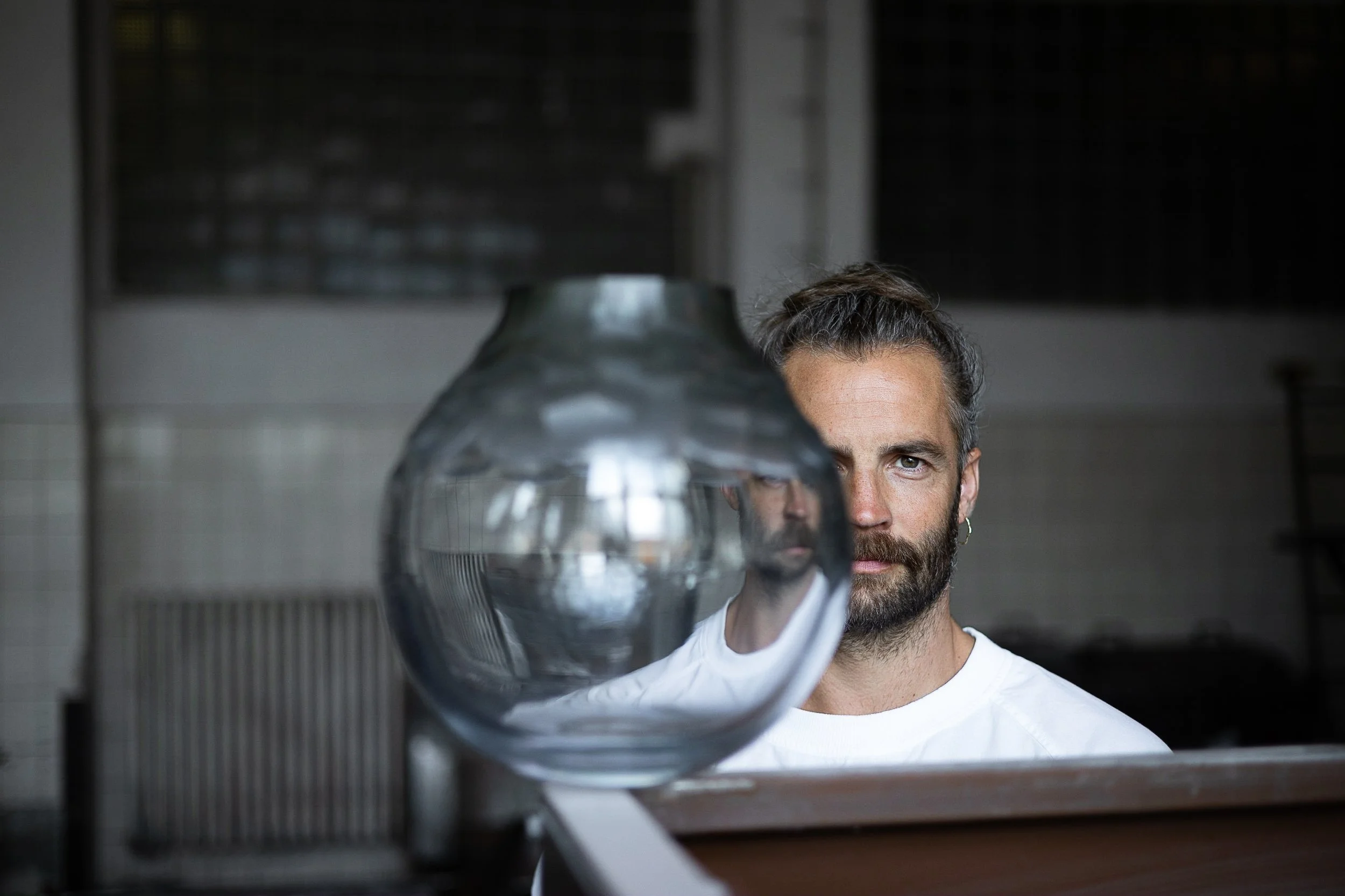 A man with a beard and tied-back hair looking at a glass object, with a reflection of his face in the glass