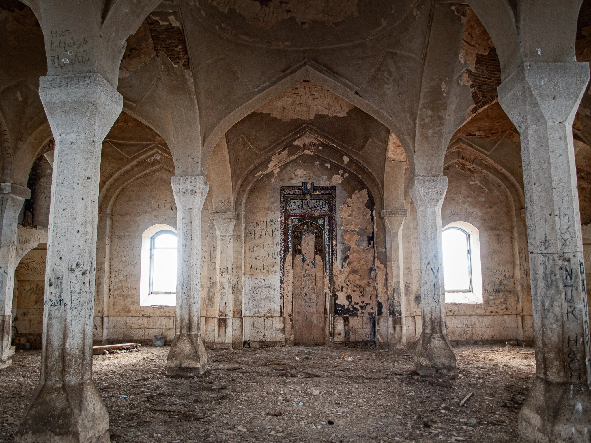 Abandoned, dilapidated room with tall arches and two small windows, graffiti on walls, debris on the ground, and a damaged wall with peeling paint.