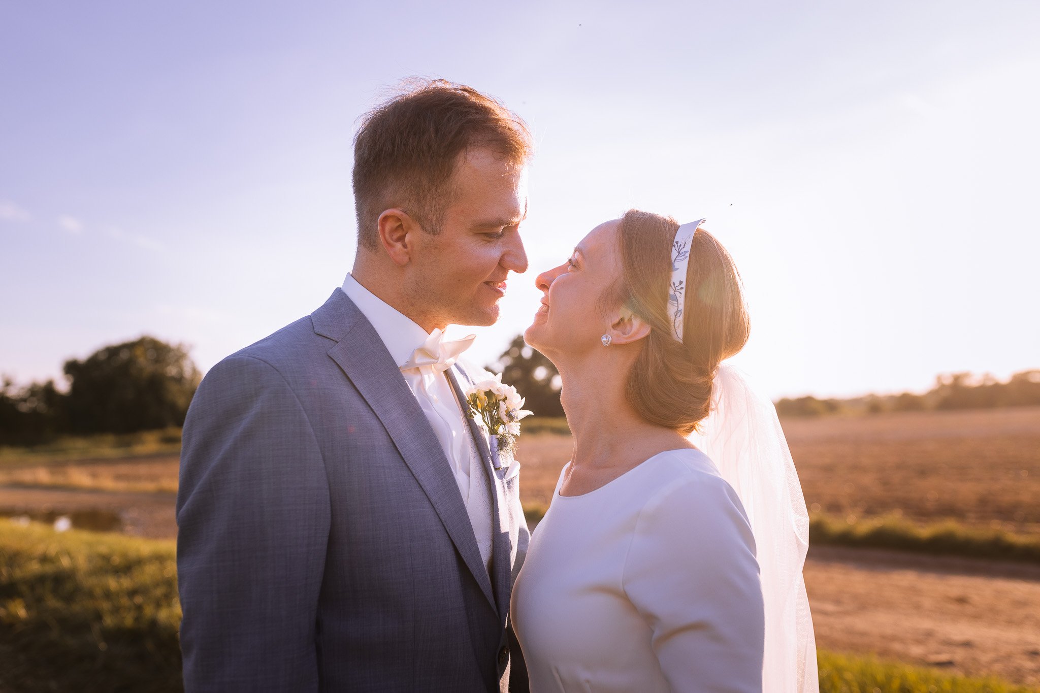 A bride and groom looking at each other and smiling outdoors during sunset, dressed in wedding attire.